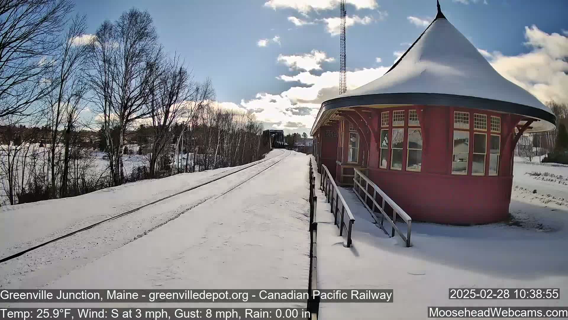 A snow-covered train platform next to a red, octagonal train station is seen on a partly sunny day with a south wind.