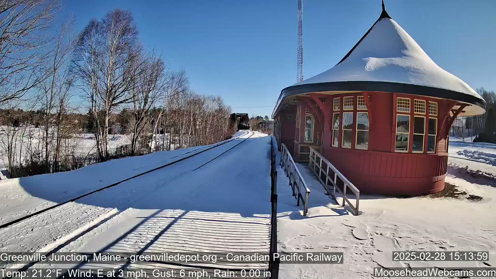 A snow-covered railroad track leads to a red, snow-dusted round train station under a clear blue sky on a sunny, cold day.