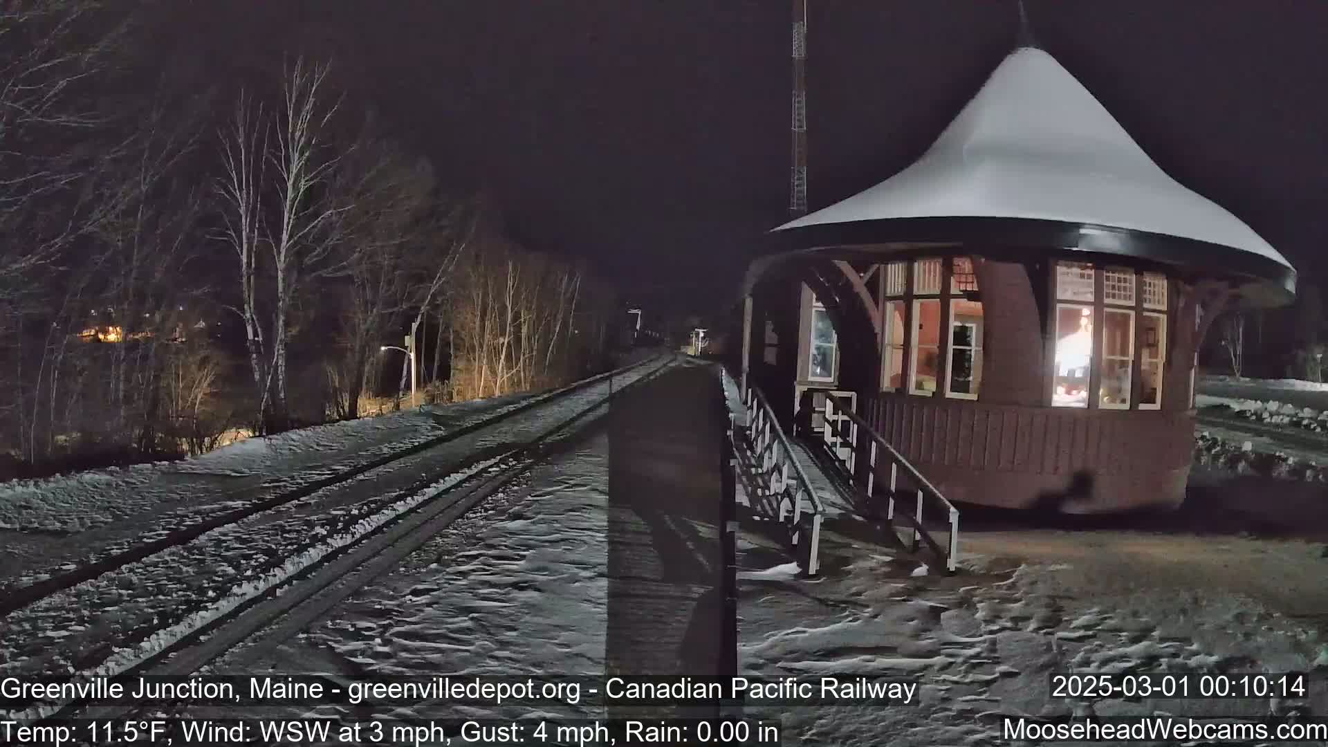 A snow-covered train platform and a lighted, snow-capped gazebo are visible at night, with a wind blowing from the west-southwest.