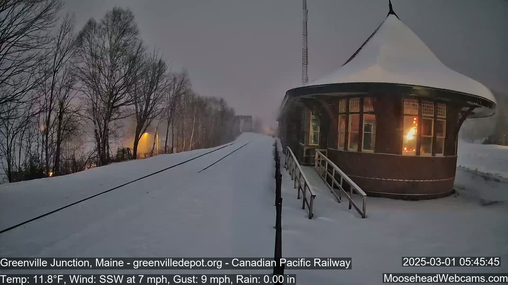 A snow-covered railway station with lights on inside is seen during a snowfall with a temperature of 11.8°F, a south-southwesterly wind at 7 mph gusting to 9 mph.