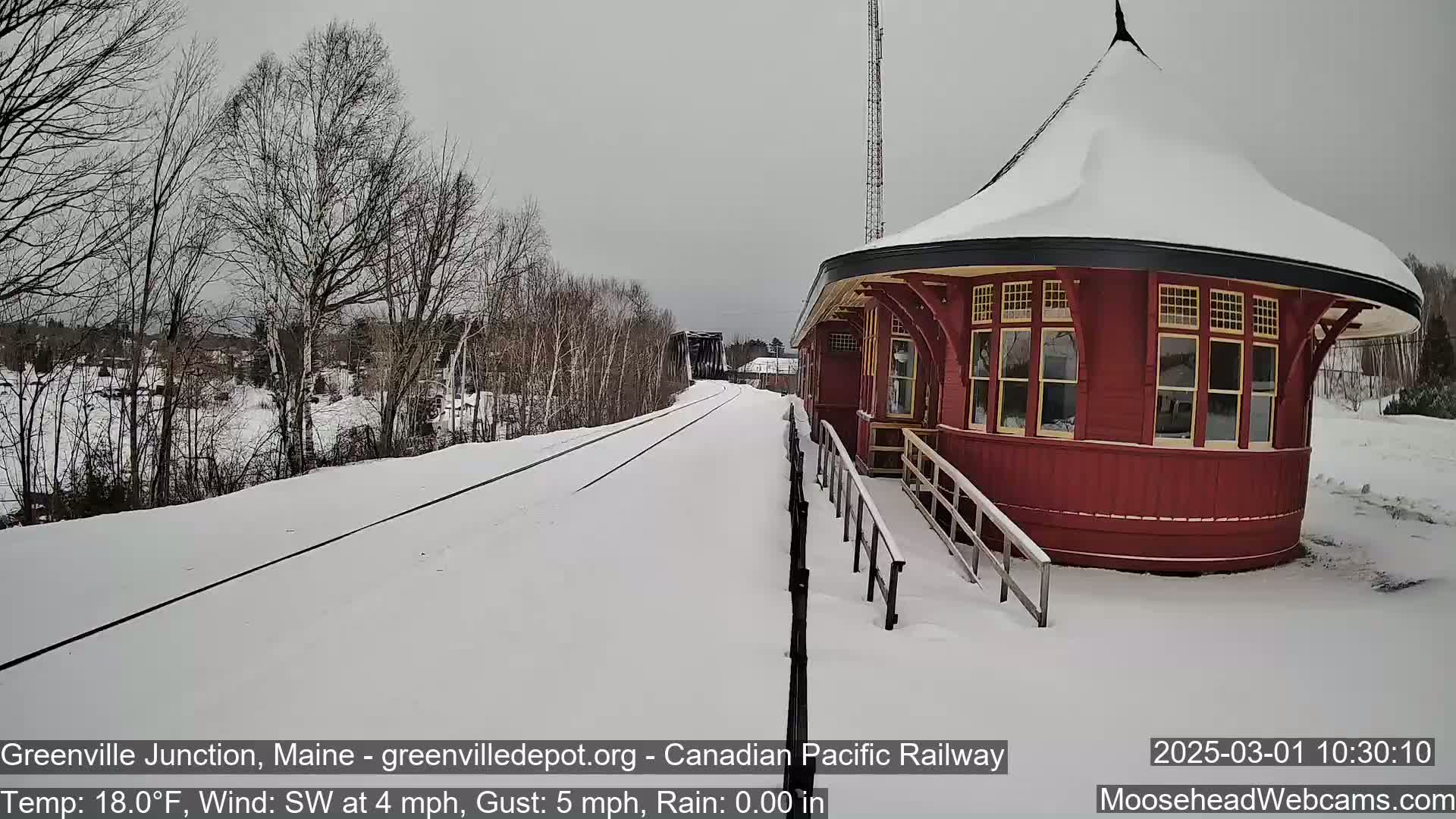 A snow-covered train platform with a red, round train station building is next to a snow-covered railway track under an overcast sky.