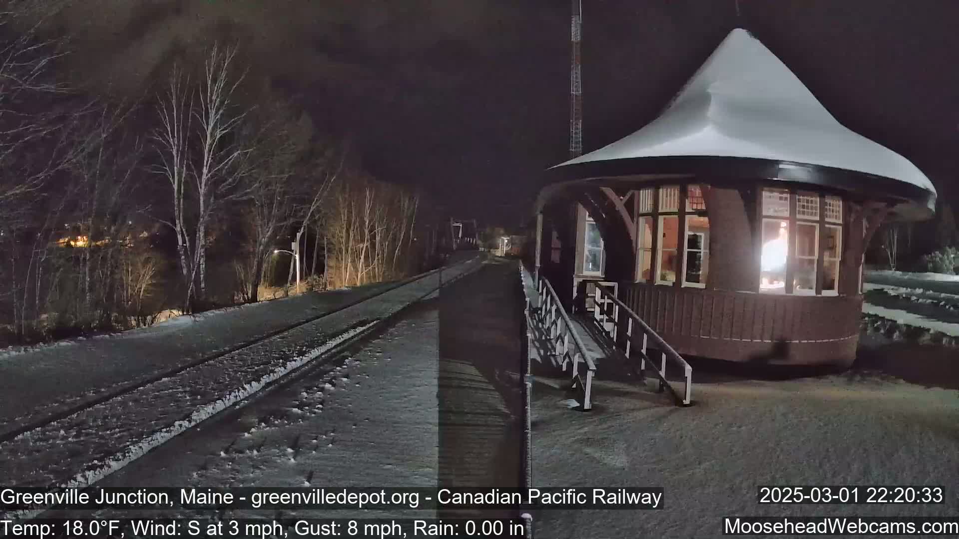 A snow-covered, dark brown train station with a snow-laden conical roof sits beside a snow-dusted railway track at night, under a clear sky with a light south wind.