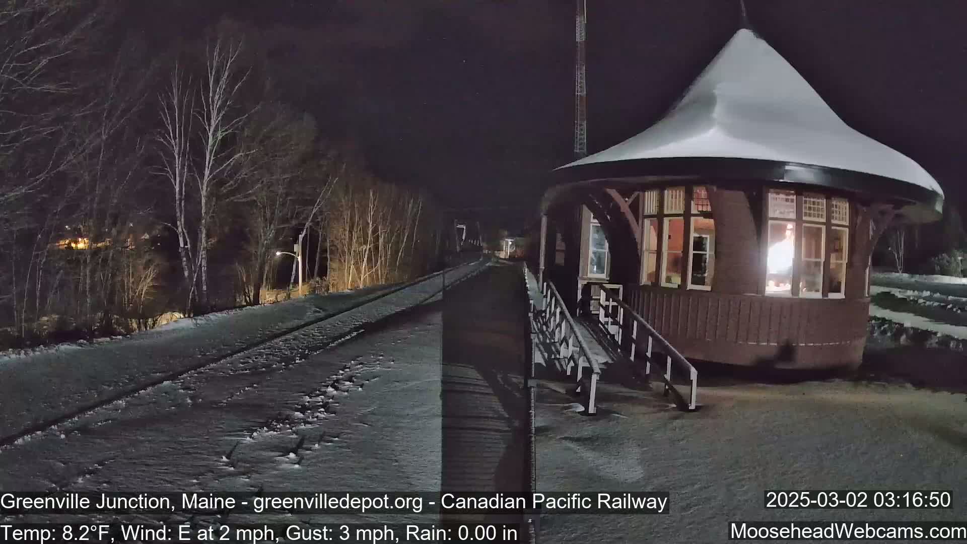 A snow-covered, round train station is illuminated at night next to a snow-covered railway track.