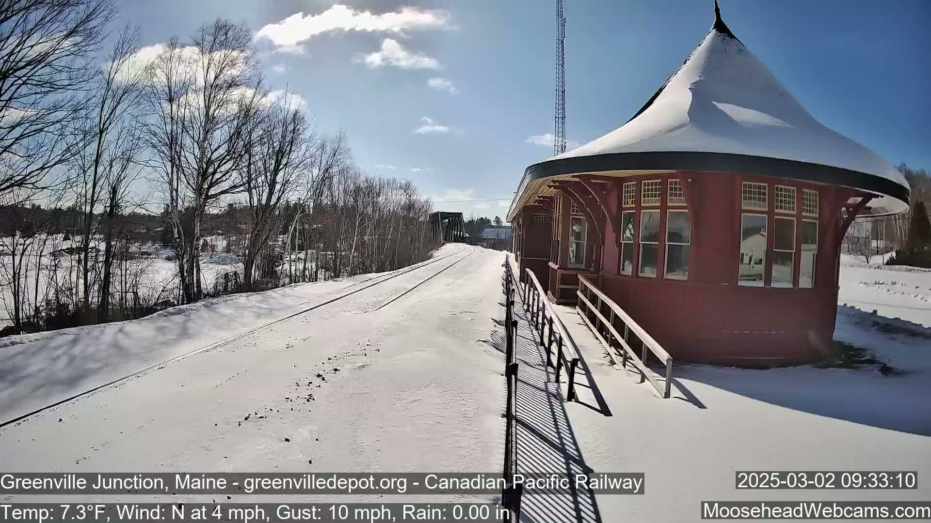 A snow-covered train platform and tracks extend toward a bridge under a partly sunny sky.