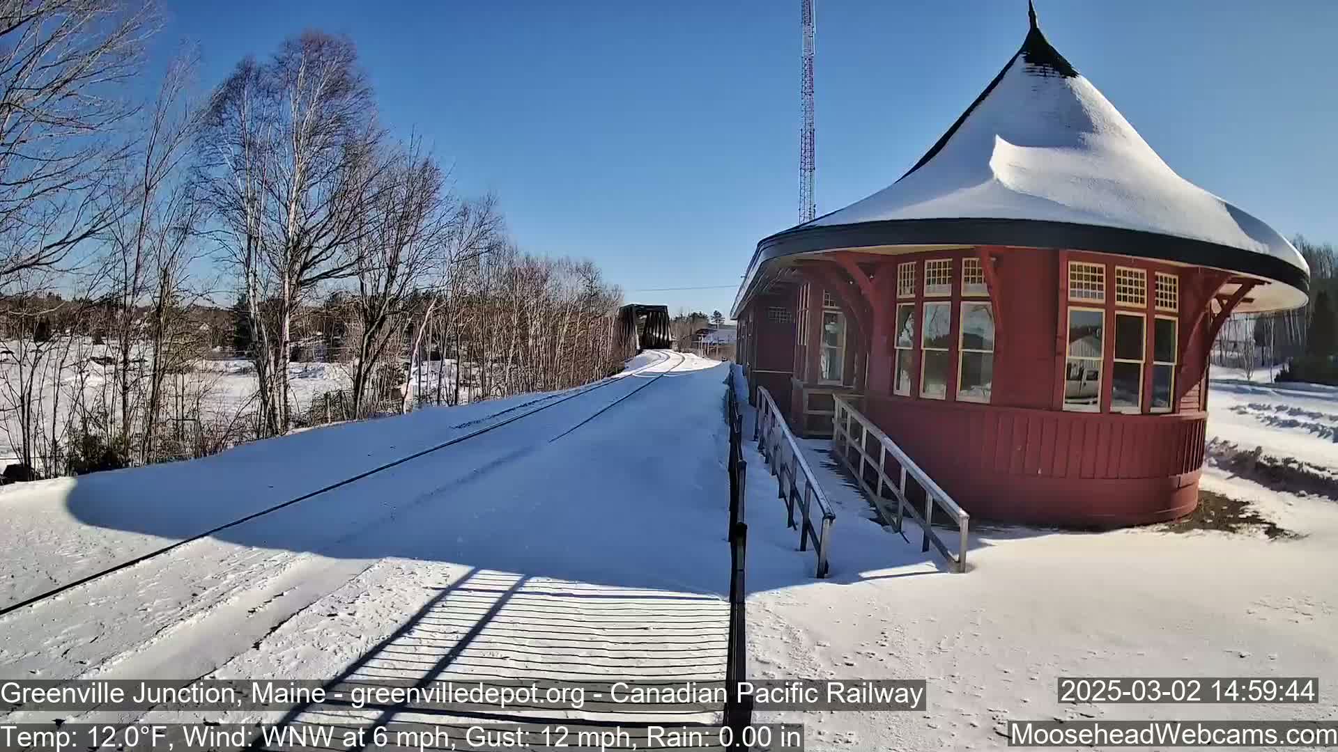 A snow-covered train platform with a red, octagonal train station and a view of train tracks leading to a bridge under a clear blue sky.