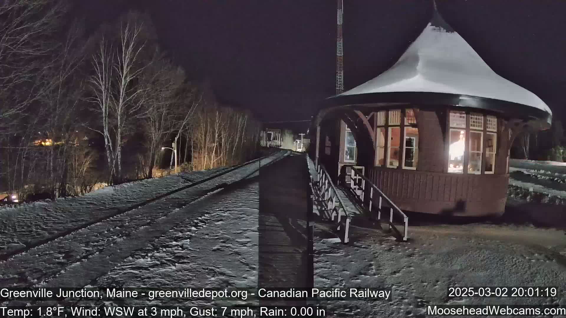 A snow-covered train platform and a round, snow-dusted building are illuminated at night, with bare trees and train tracks in the background.