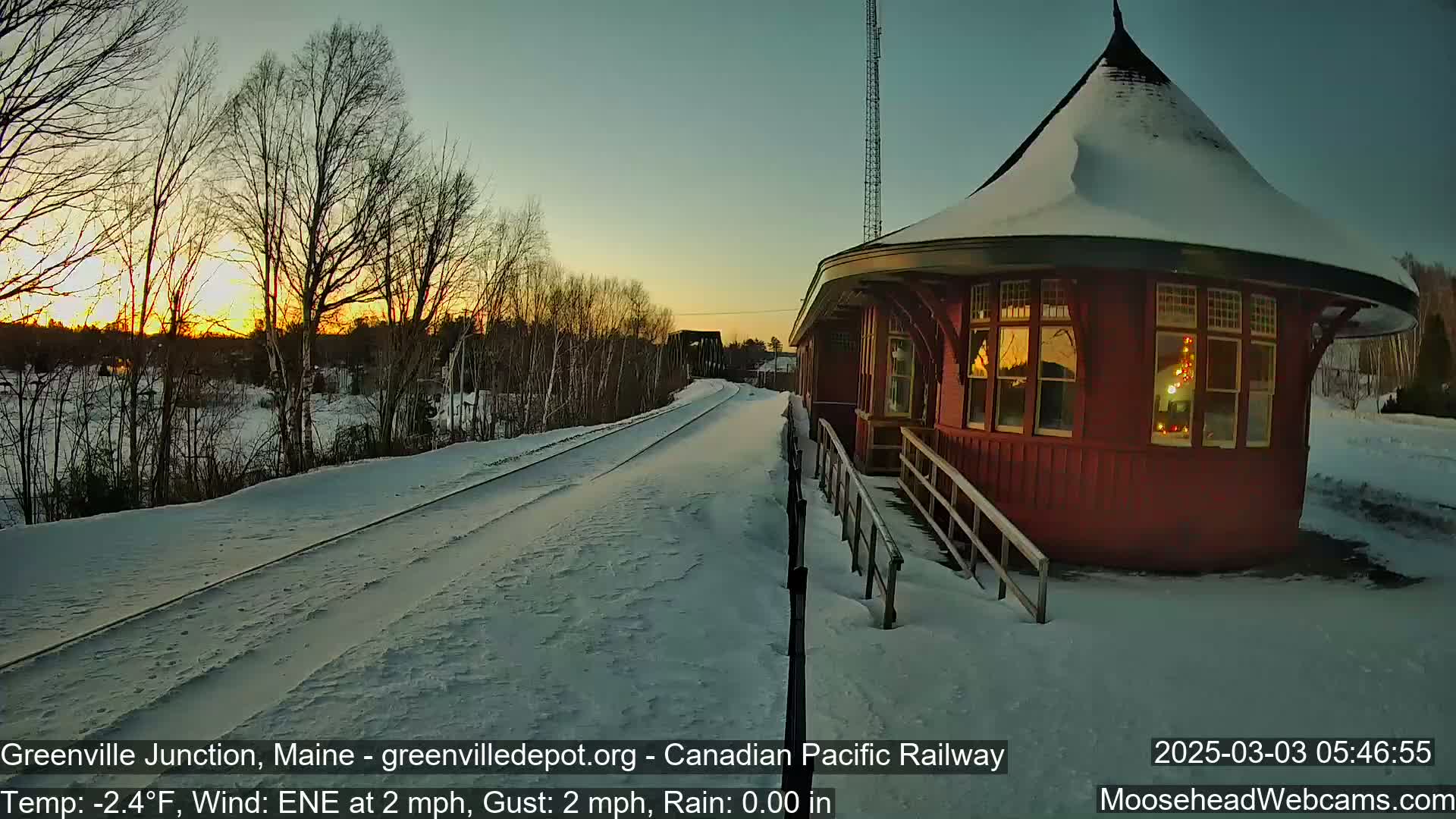 A snow-covered train platform with a red, round building is illuminated by a setting sun, with a light wind.
