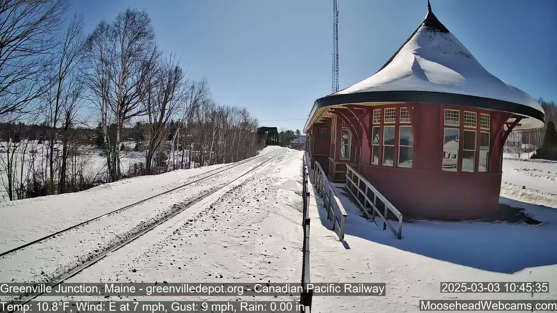A snow-covered train station and railway tracks extend into the distance under a bright, sunny sky.