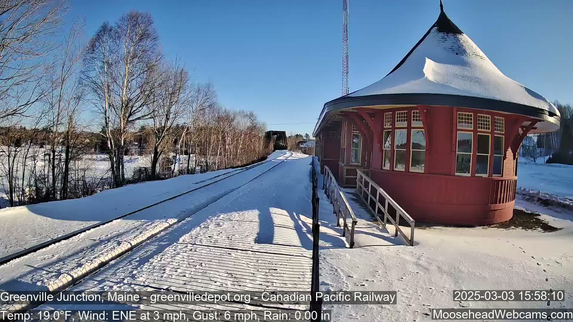 A snow-covered train platform with a red, round train station and tracks extending to a bridge under a clear blue sky.