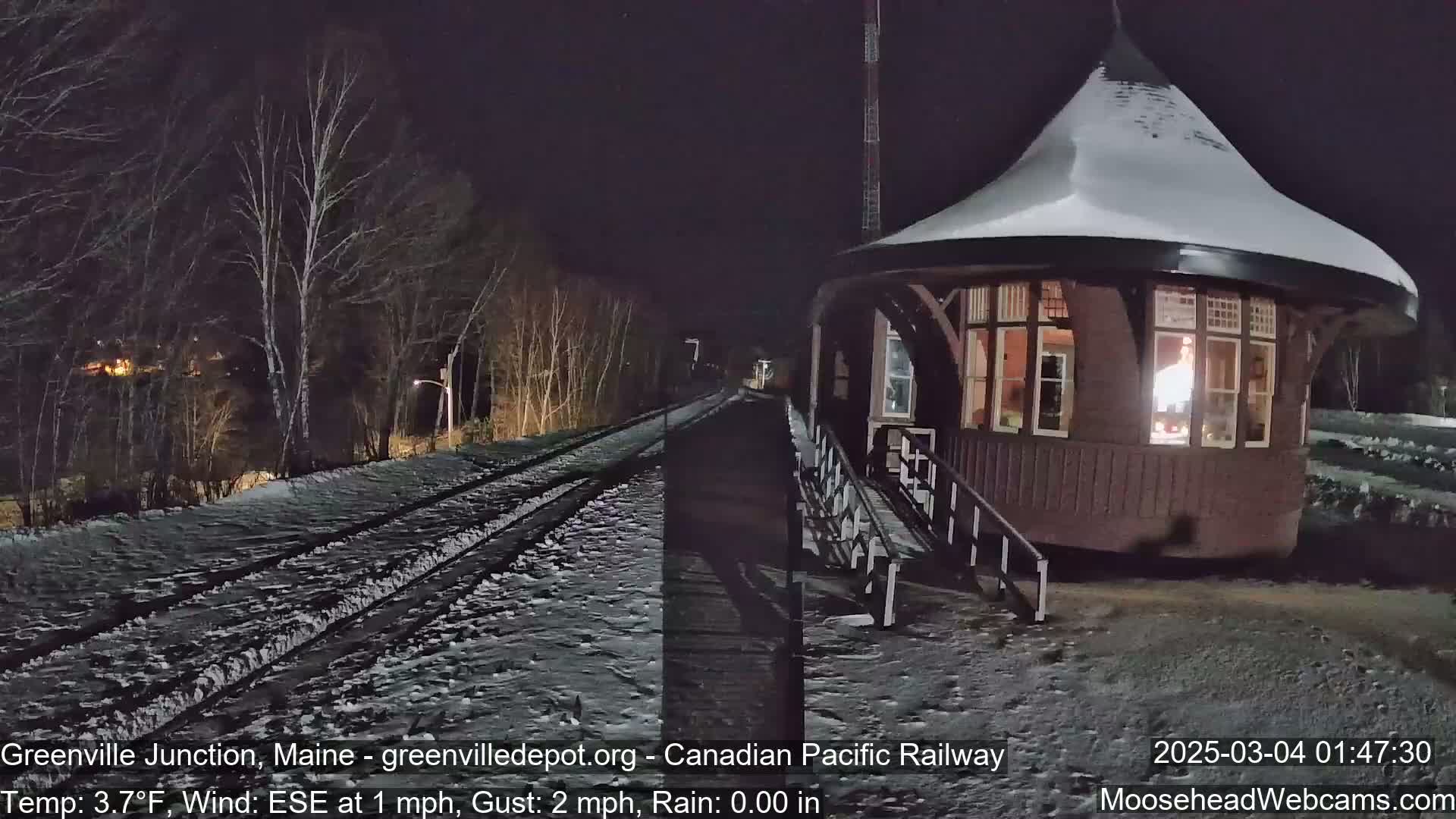 A snow-covered train platform with a small, round building is illuminated at night by a light inside, with bare trees and a railroad track in the background.