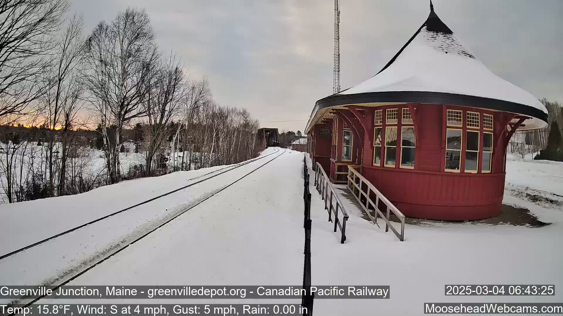 A snow-covered railway track leads to a red, round train station under a partly cloudy sky.