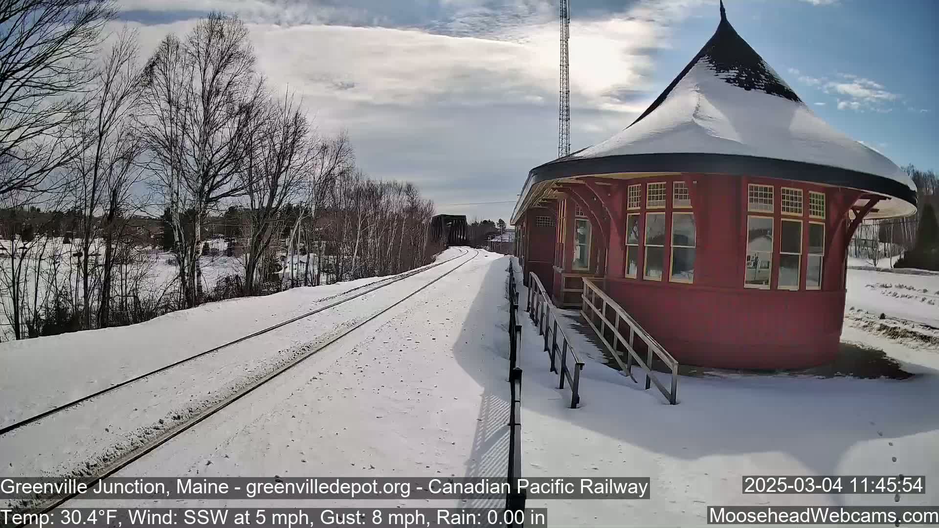 A snow-covered train platform with a red octagonal building sits beside train tracks extending into a winter landscape under a partly sunny sky.