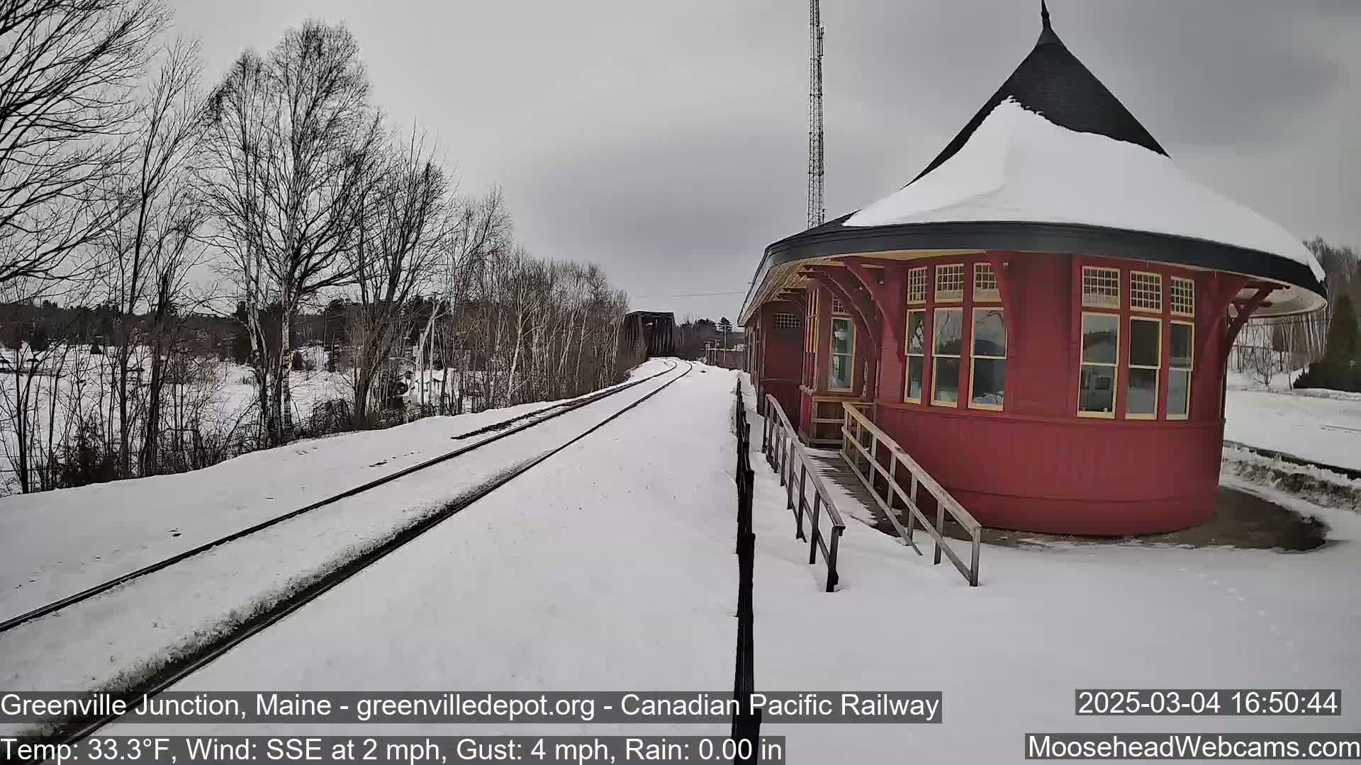 A snow-covered train platform with a red, round train station and railway tracks extends into the distance under an overcast sky.