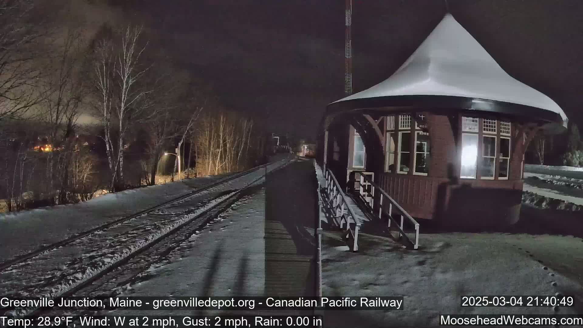 A snow-covered train station platform at night, with a wooden gazebo-like structure and train tracks leading into a dark, snow-covered landscape under a mostly clear sky.