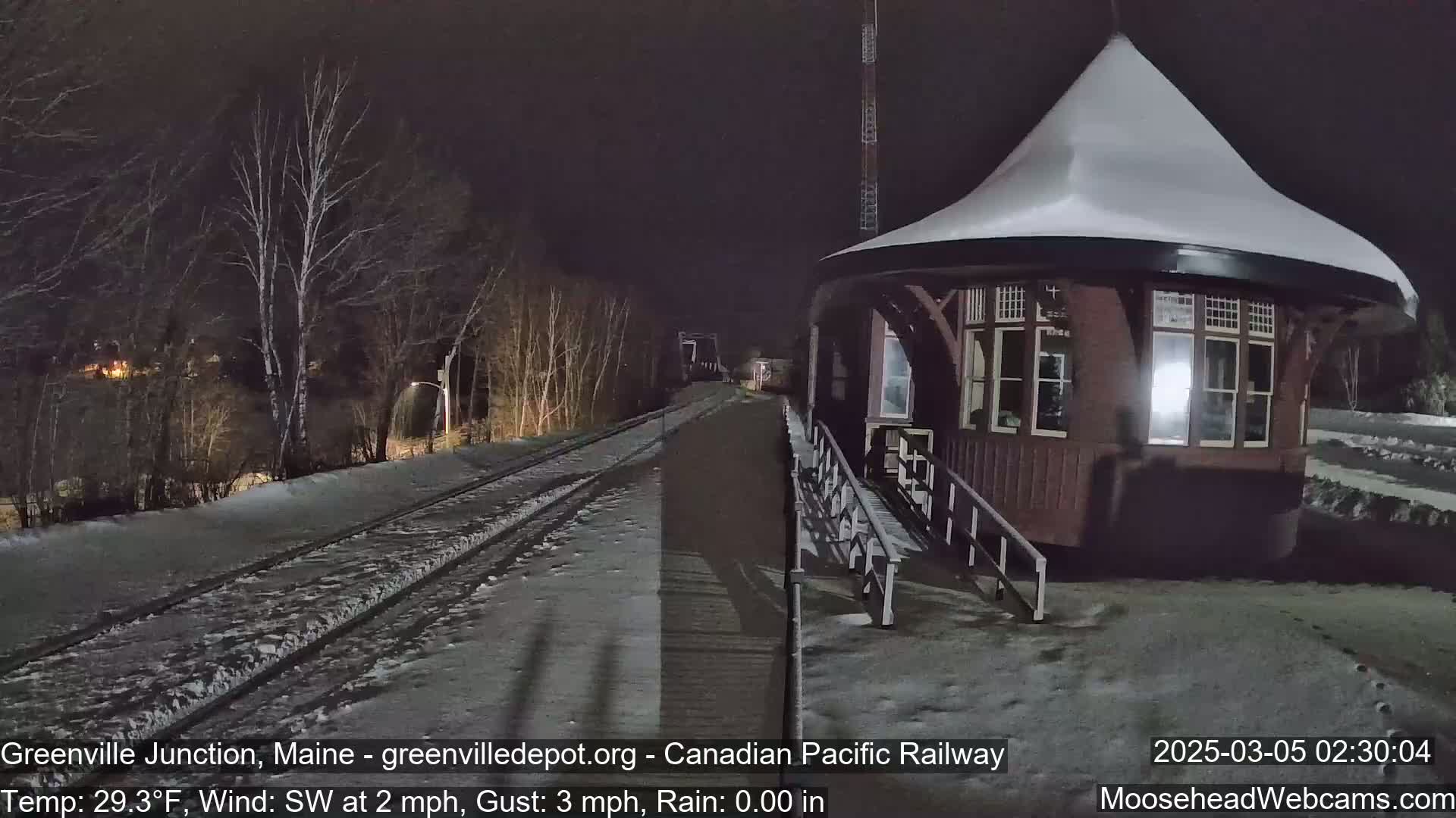 A snow-covered, dark red train station stands beside a snow-dusted railway track at night under a dark sky.