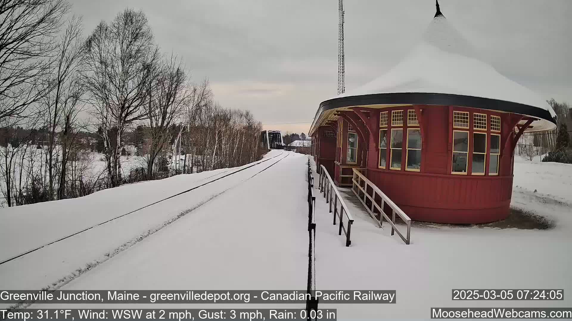 A snow-covered train station and railway track are visible in the lightly snowing scene.