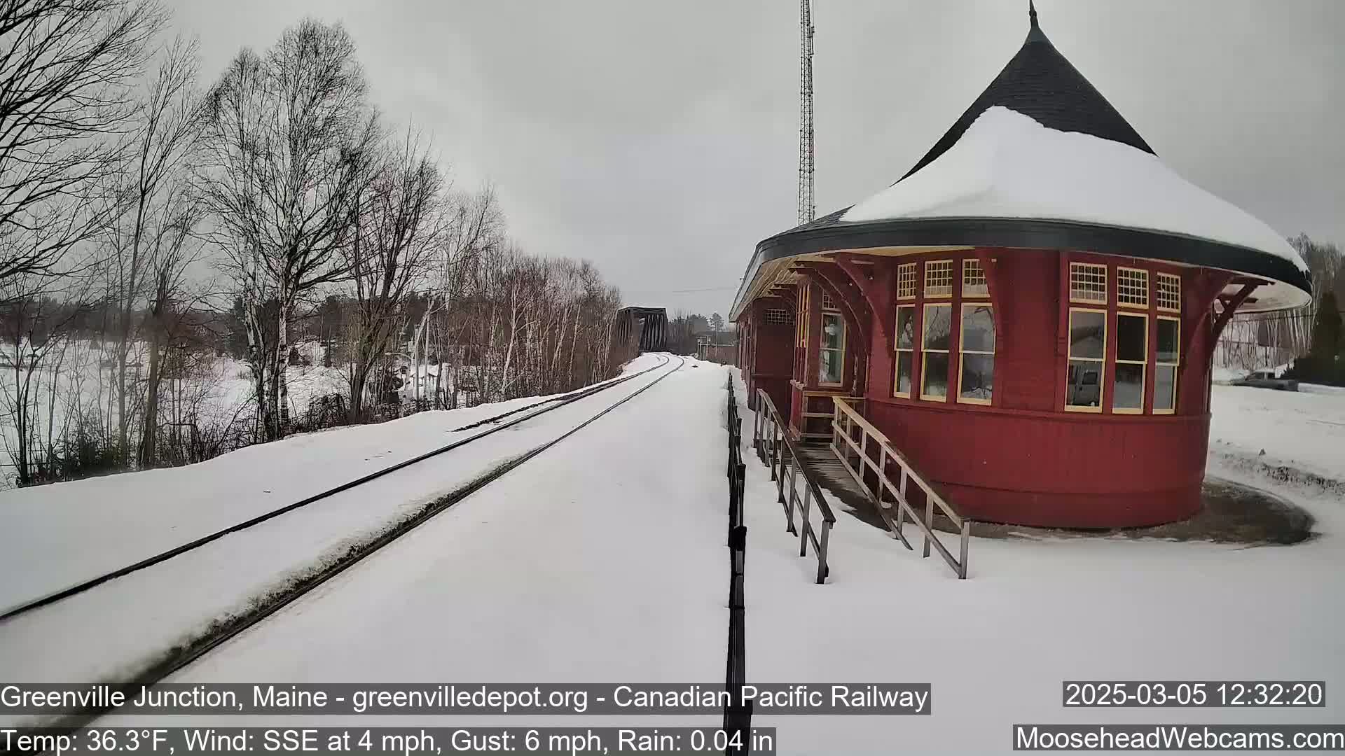 A snow-covered train platform with a red, round train station and a view of snow-covered tracks extending to a bridge in the distance under an overcast sky.