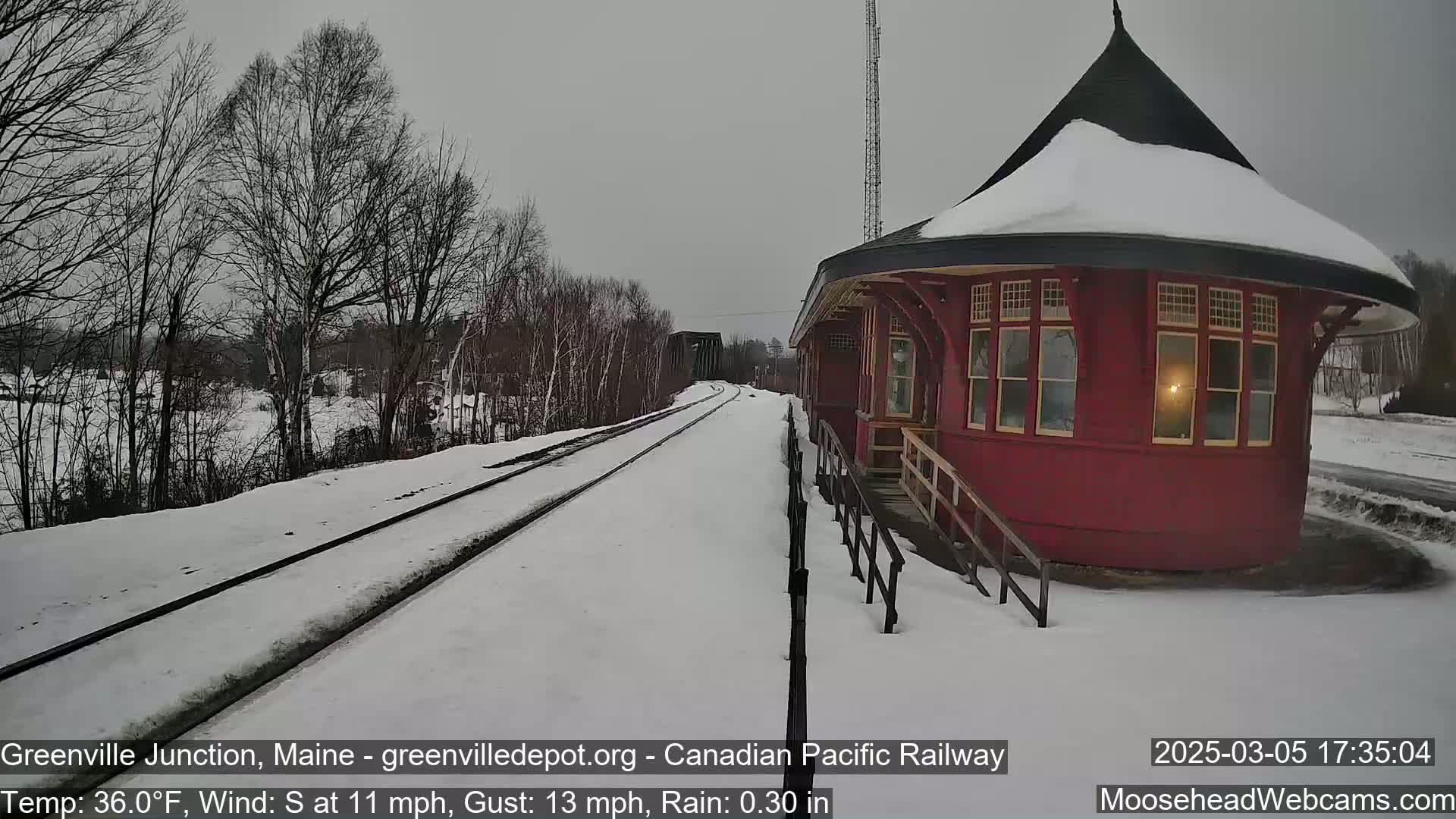 A snow-covered train track leads to a red, round train station under a gray sky with a south wind and light rain.
