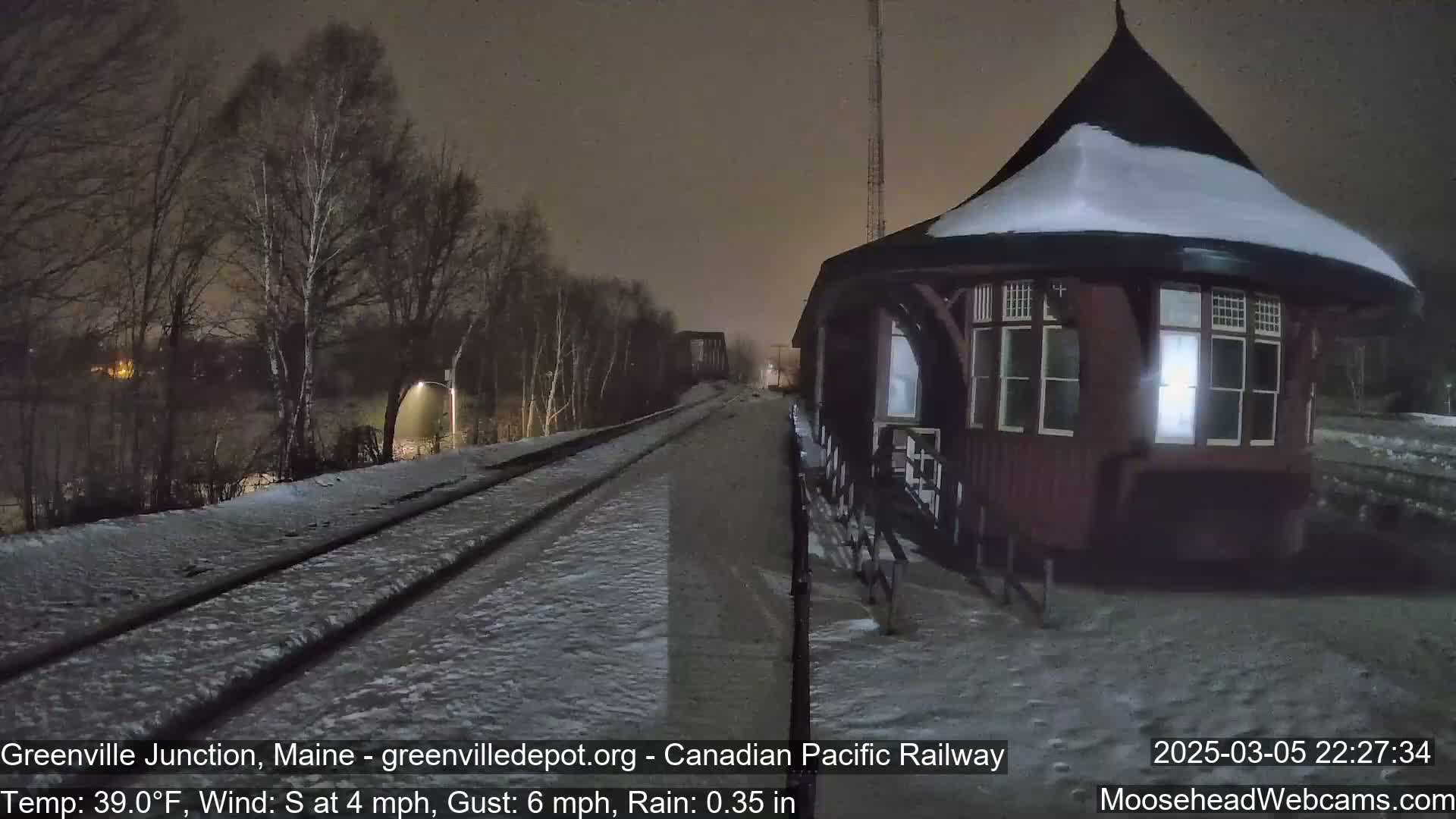 A snow-covered train platform and tracks are visible at night next to a dark-red, snow-dusted train station under rainy conditions.