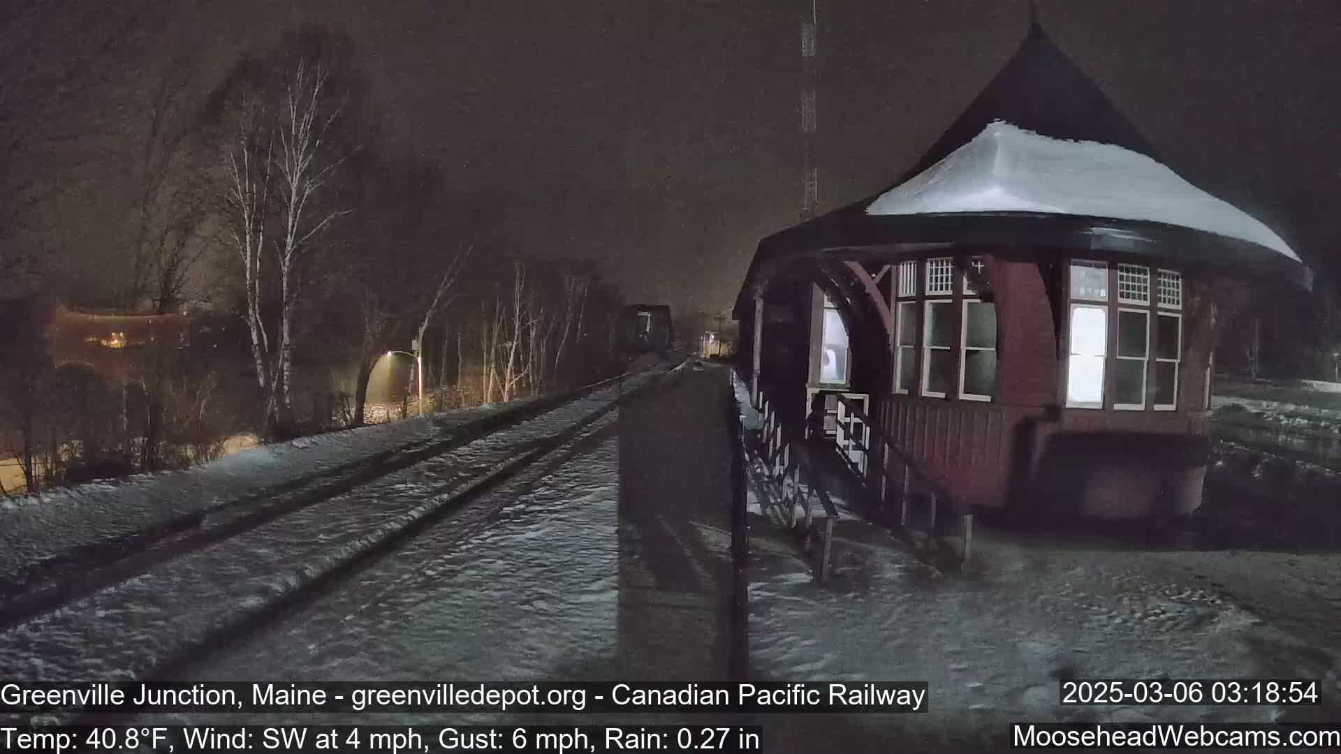 A snow-covered, dark-red octagonal train station stands next to railway tracks in the rain at night.