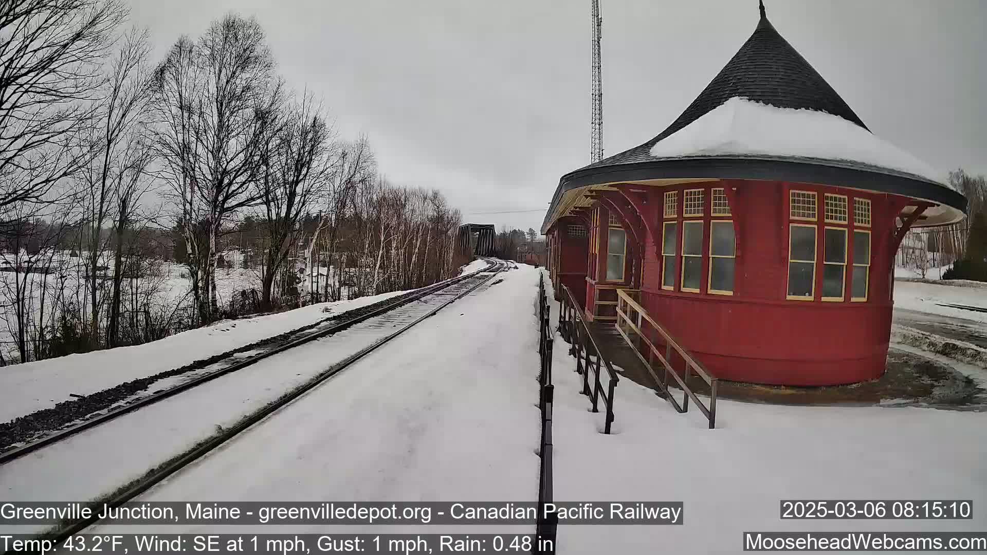 A snow-covered railroad track leads to a red octagonal train station under a gray sky, with light rain.