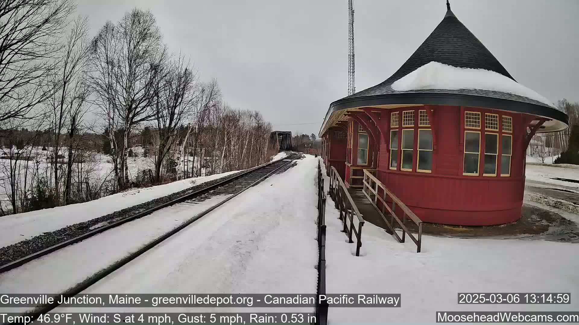 A red, round train station sits beside snow-covered train tracks on an overcast day with a light south wind and rain.