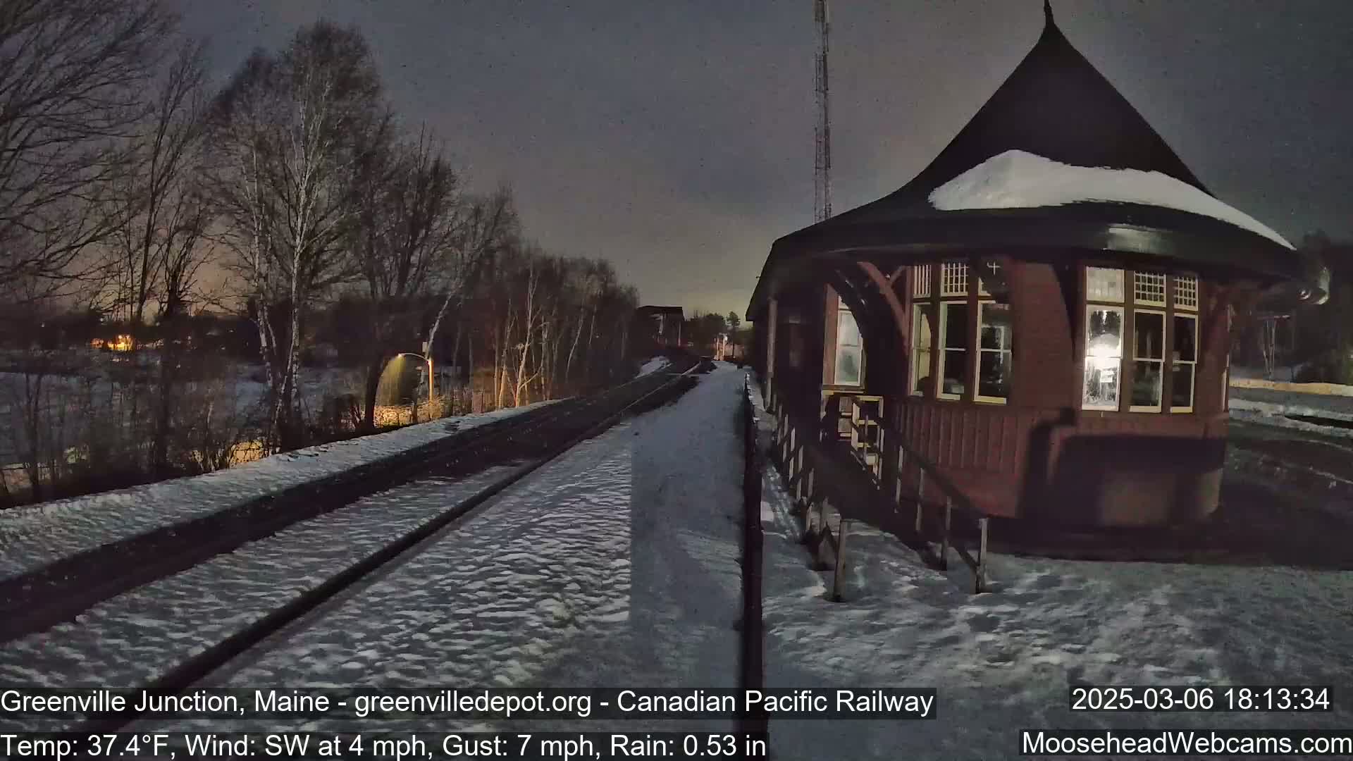 A snow-covered railway track leads to a dark red octagonal train station under a nighttime sky with light rain.