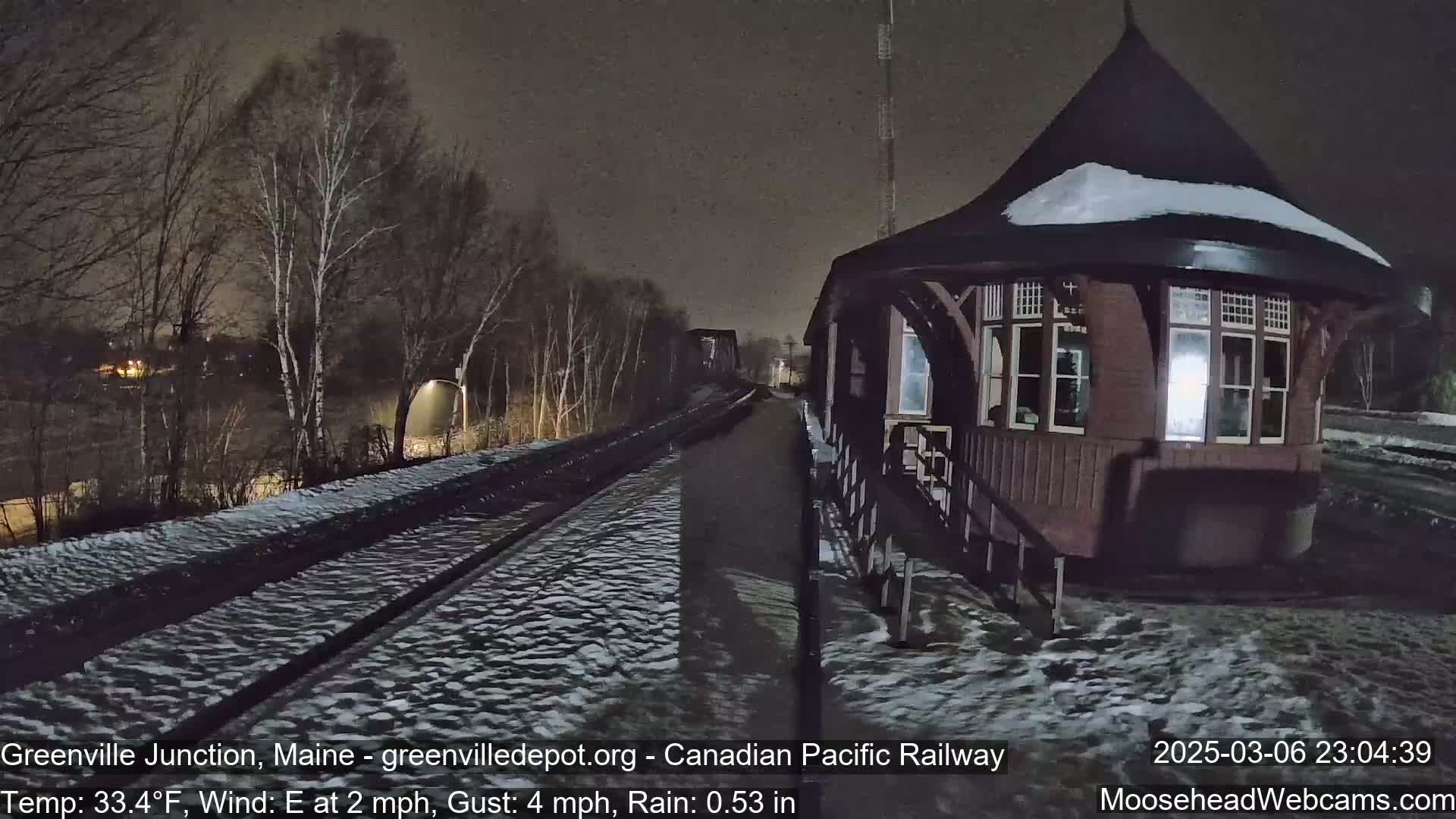 A snow-covered railroad track extends toward a dark, wooden, octagonal train station under a nighttime sky with light rain.