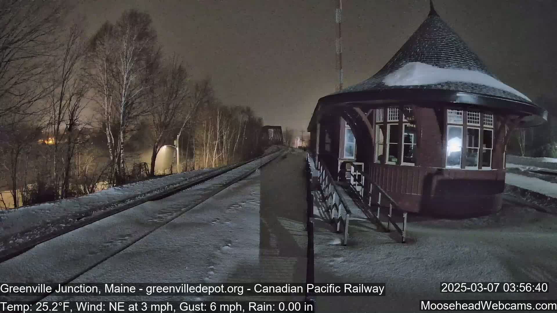 A snow-covered train platform with a dark brown, conical-roofed building is illuminated at night by a nearby streetlight.