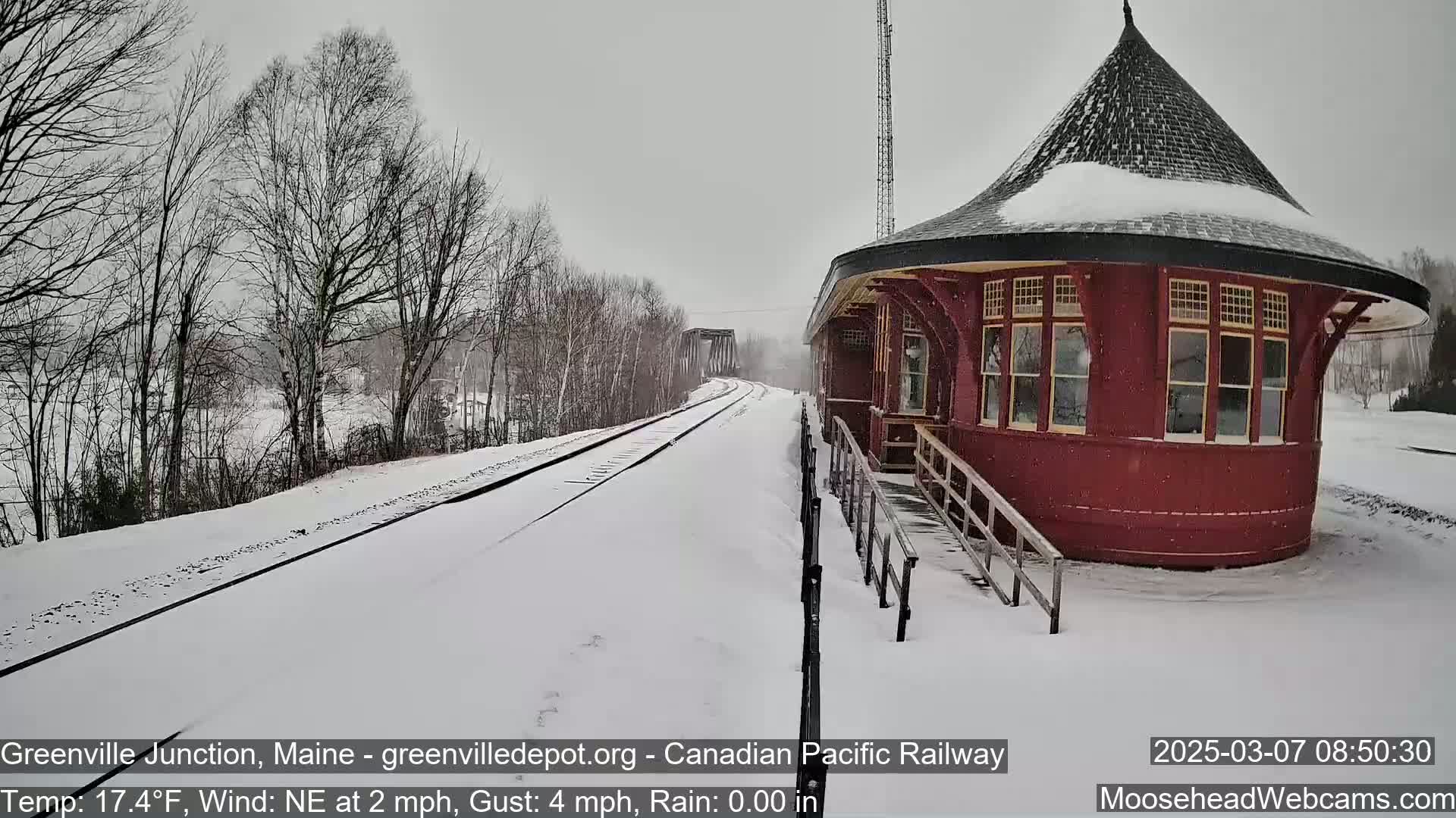 Snow falls on a snow-covered train platform next to a round, red train station and railway tracks.