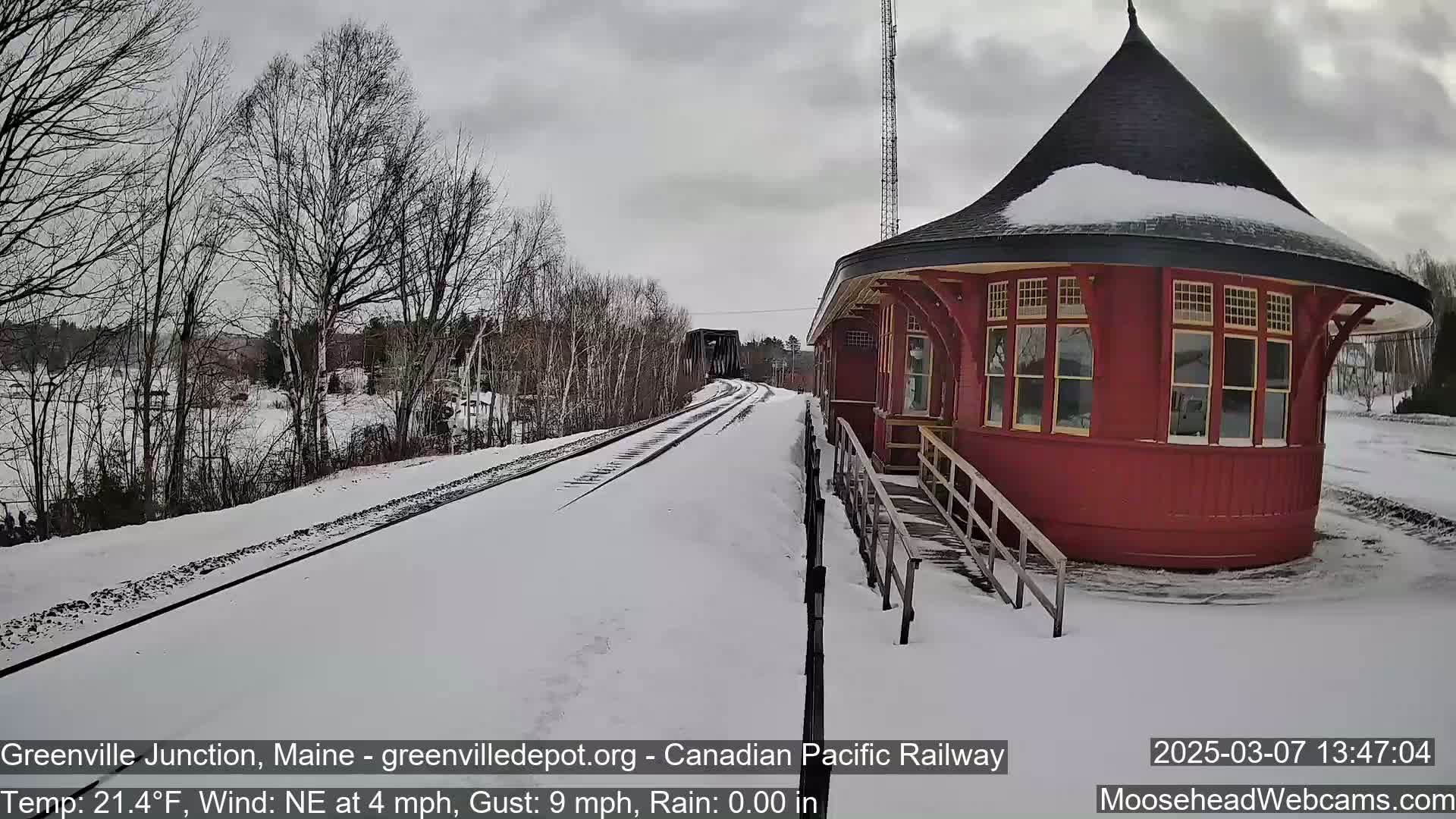 A snow-covered train track leads to a red, round train depot under a cloudy sky.