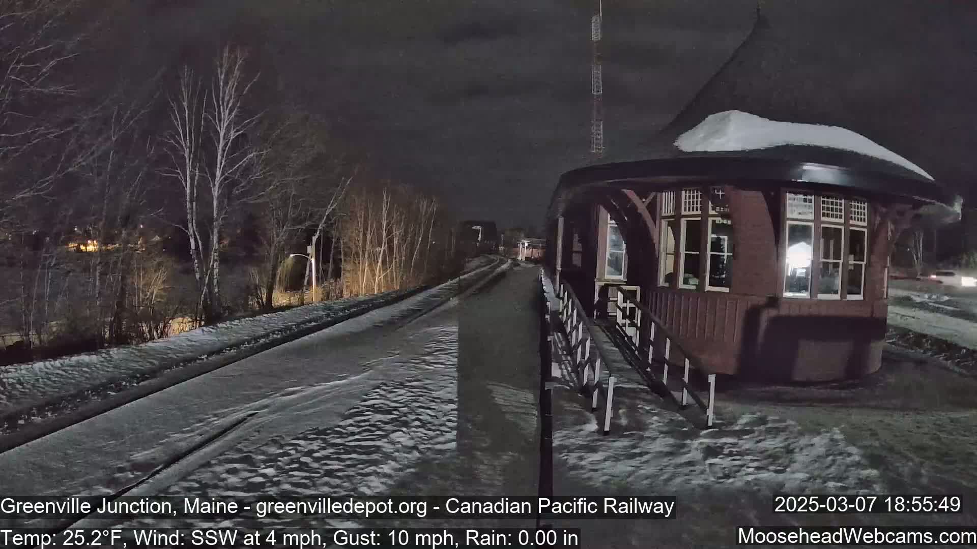 A snow-covered road leads to a dark-brown, octagonal train station at night under a dark sky with a light rain.