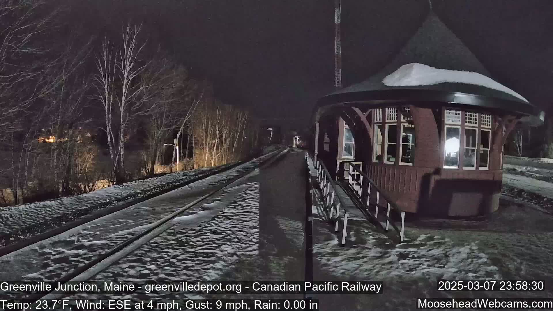 A snow-covered train platform and tracks are visible at night, with a dark-red, octagonal train station building to the right, under a dark sky with a wind from the east-southeast at 4 mph and gusts up to 9 mph.