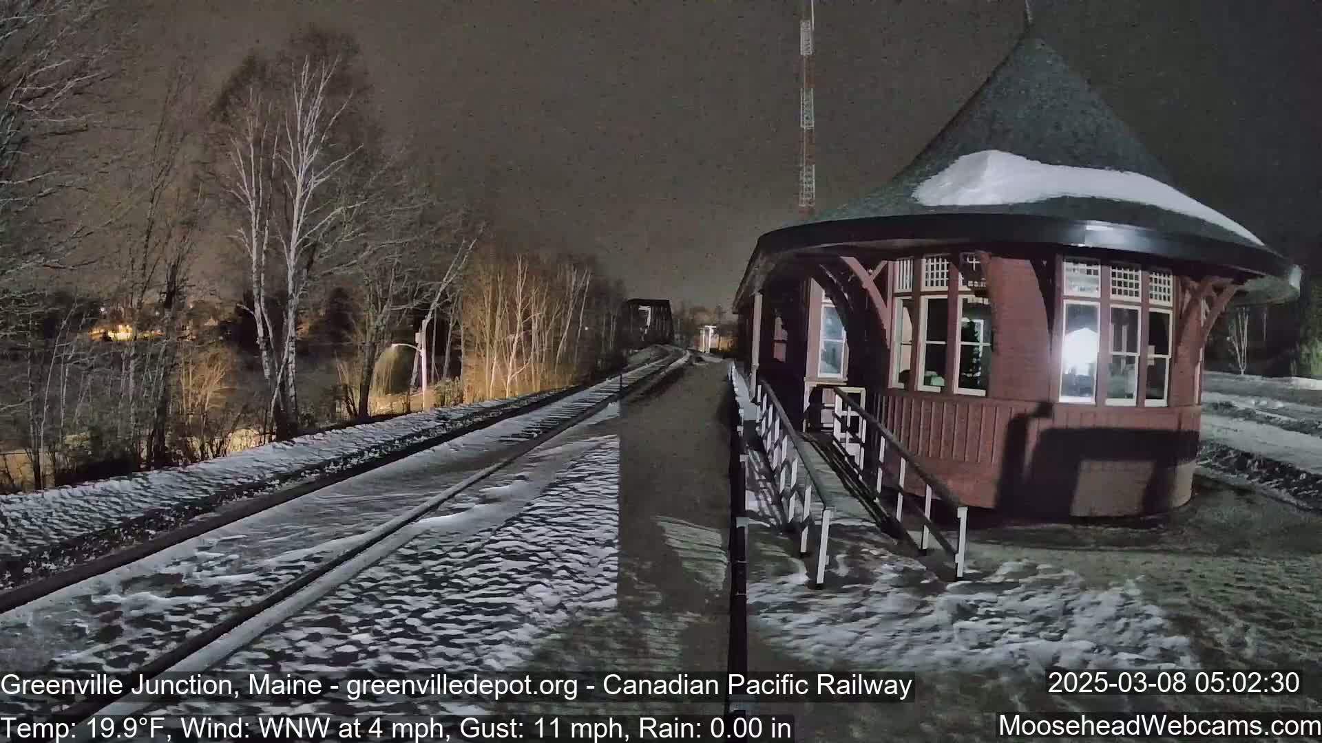 A snow-covered train platform and a red octagonal train station are visible at night under a dark sky.
