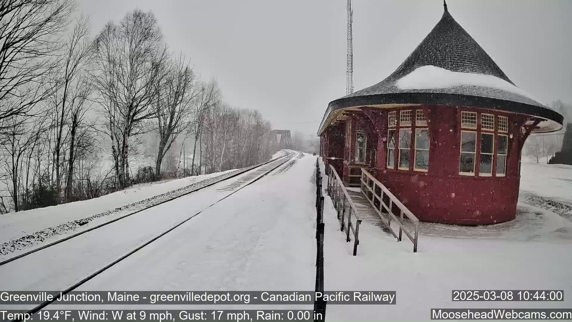 Snow falls on a snow-covered railway track leading to a red, round train depot.