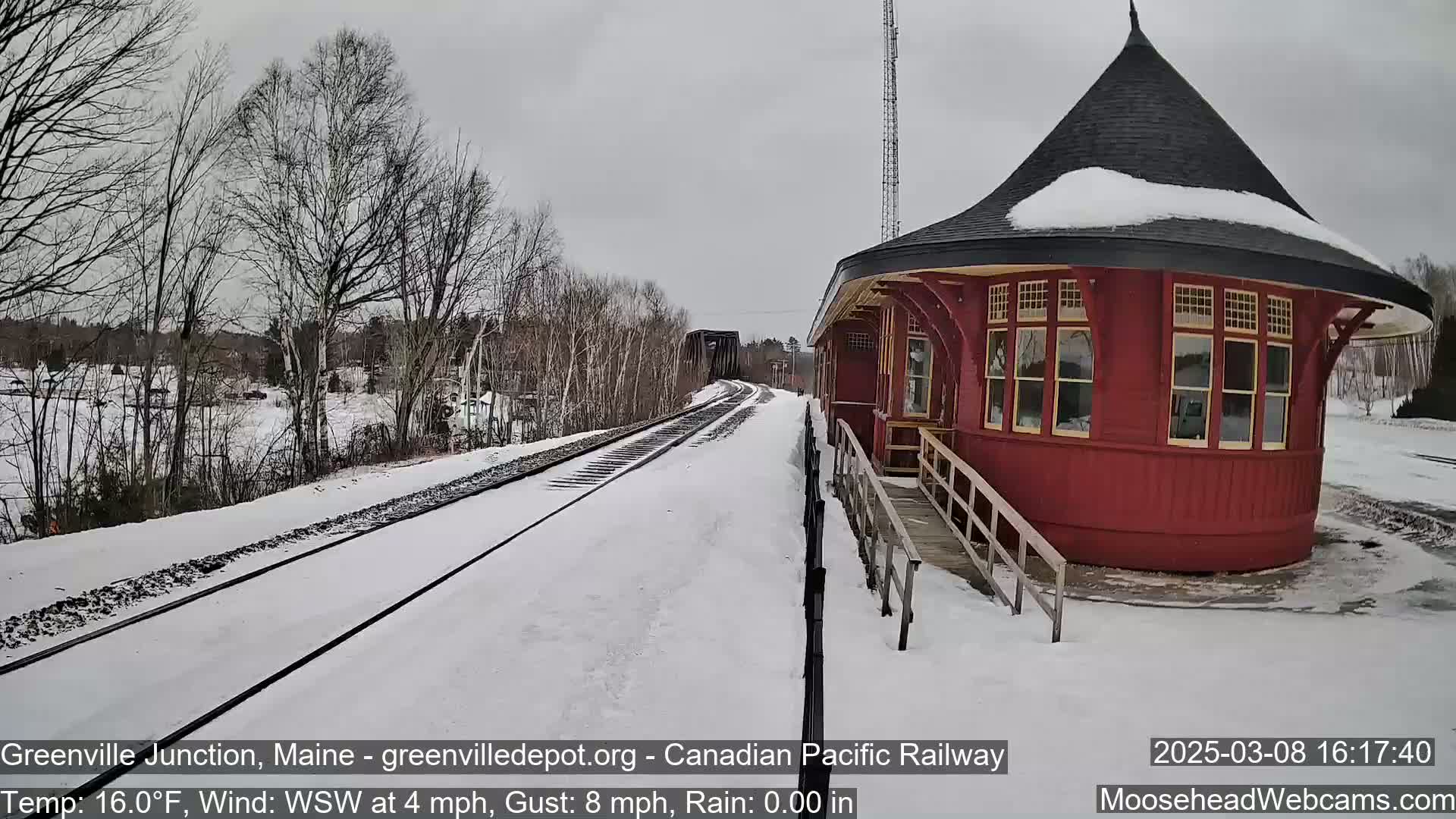 A red, octagonal train station stands beside snow-covered train tracks on a cloudy day.