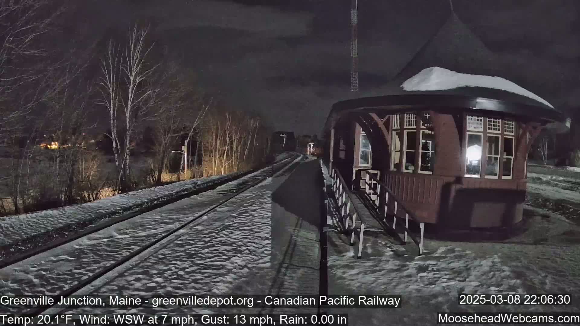 A snow-covered train platform and tracks are visible at night, with a dark brown octagonal building adjacent to the tracks, under a dark sky with a WSW wind at 7 mph and gusts up to 13 mph.