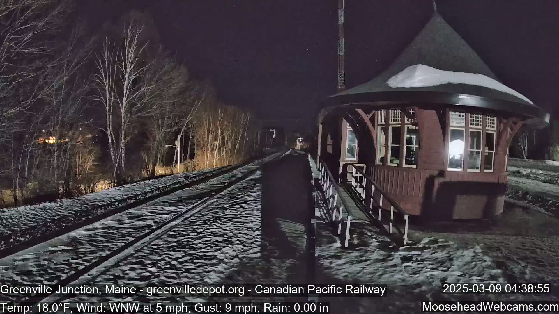 A snow-covered train platform and tracks are visible at night under a dark sky, with a dark-red wooden train station building in the foreground.