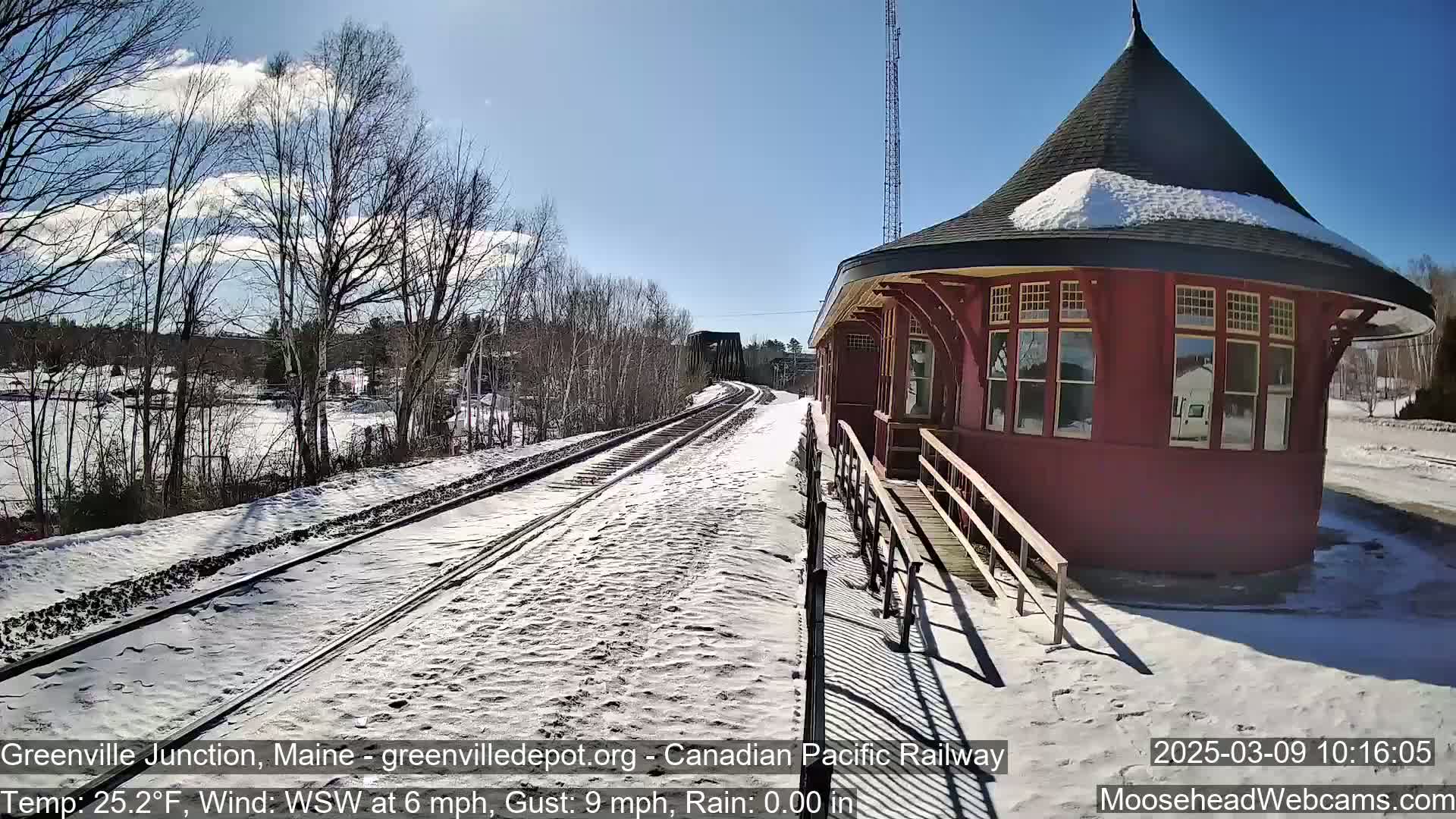 A snow-covered train platform and tracks extend towards a red, octagonal train station under a sunny sky.