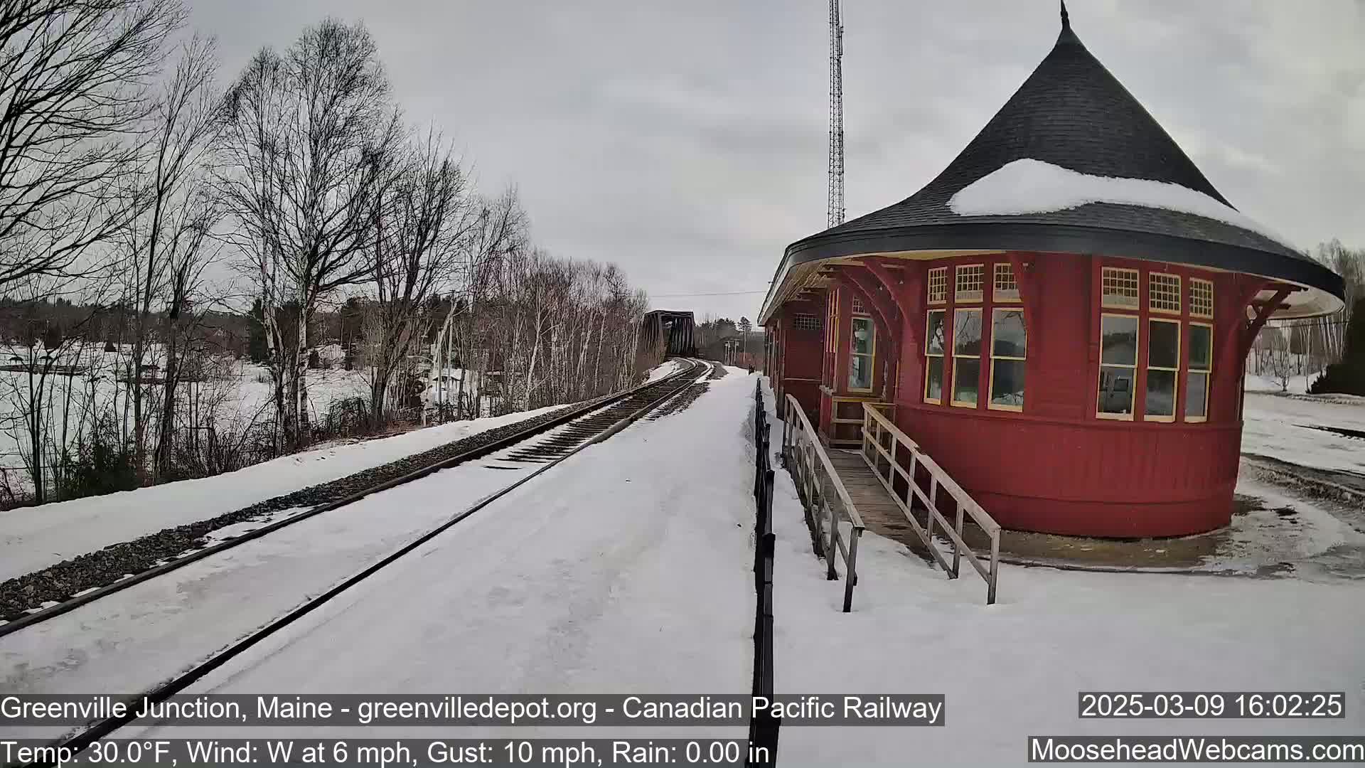 A snow-covered railway track leads to a red, octagonal train station under a cloudy sky.