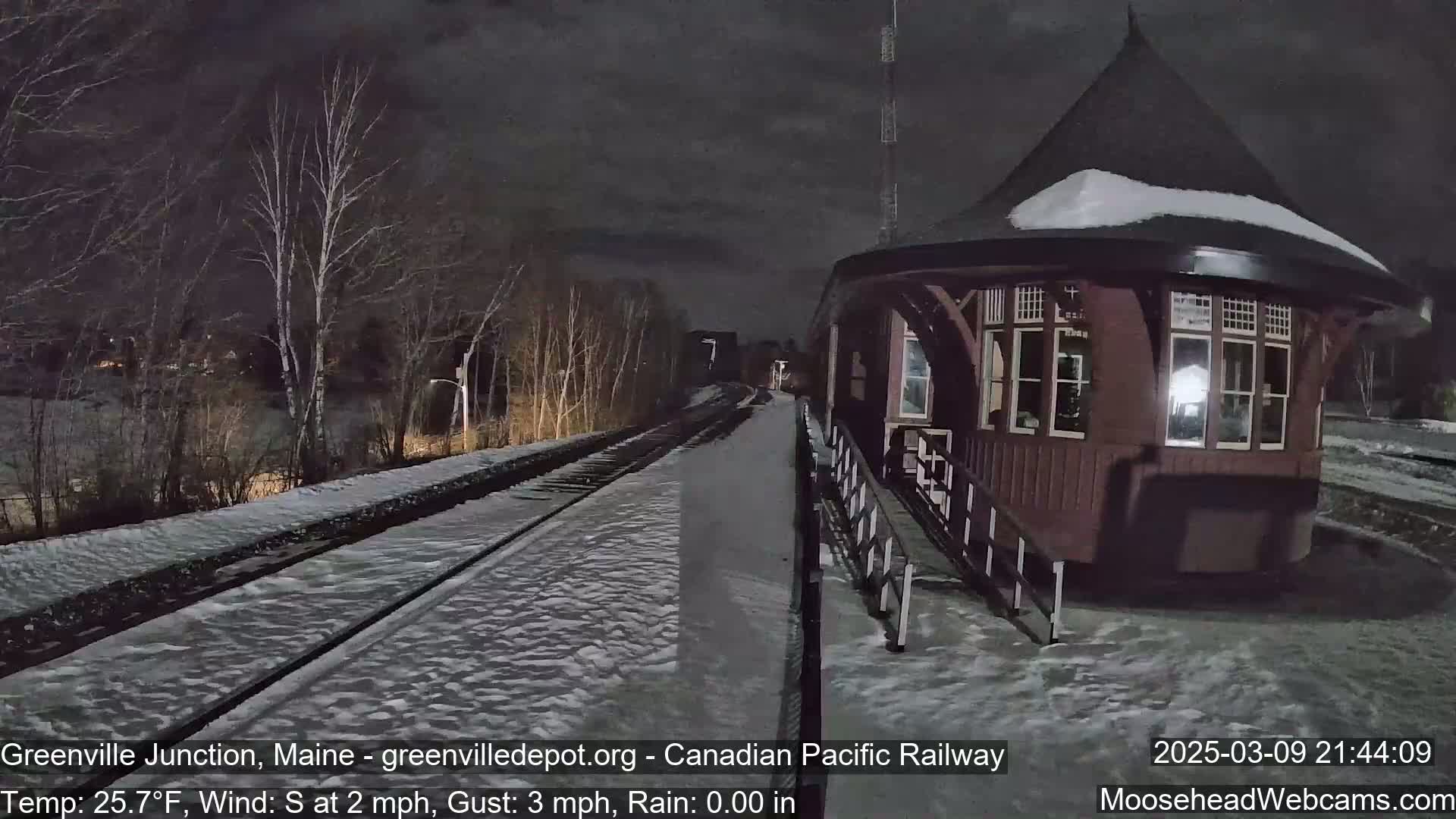 A snow-covered train track leads to a dark brown octagonal train station under a nighttime sky.