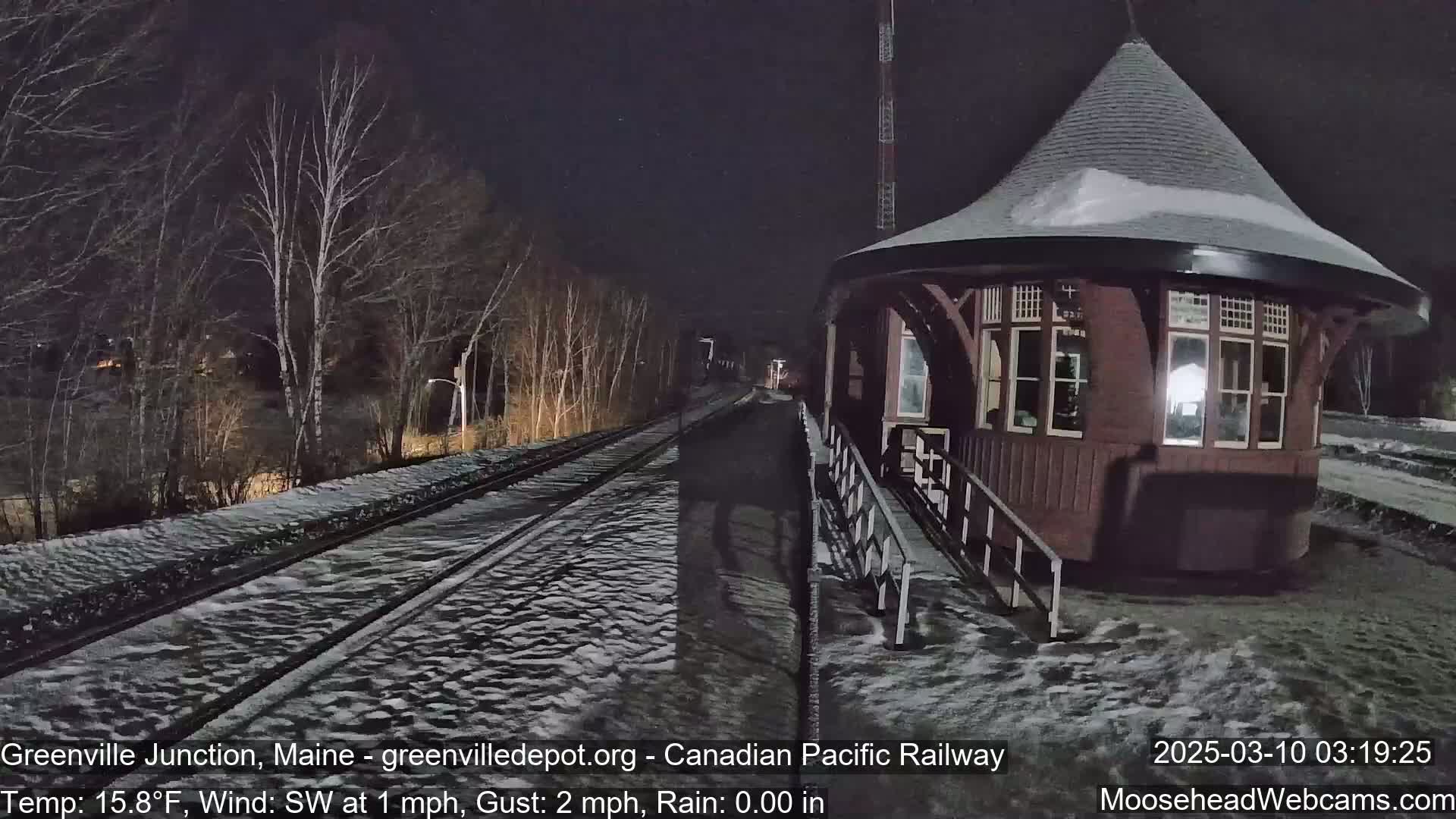 A snow-covered train platform with a dark brown octagonal building sits next to a set of train tracks under a nighttime sky.