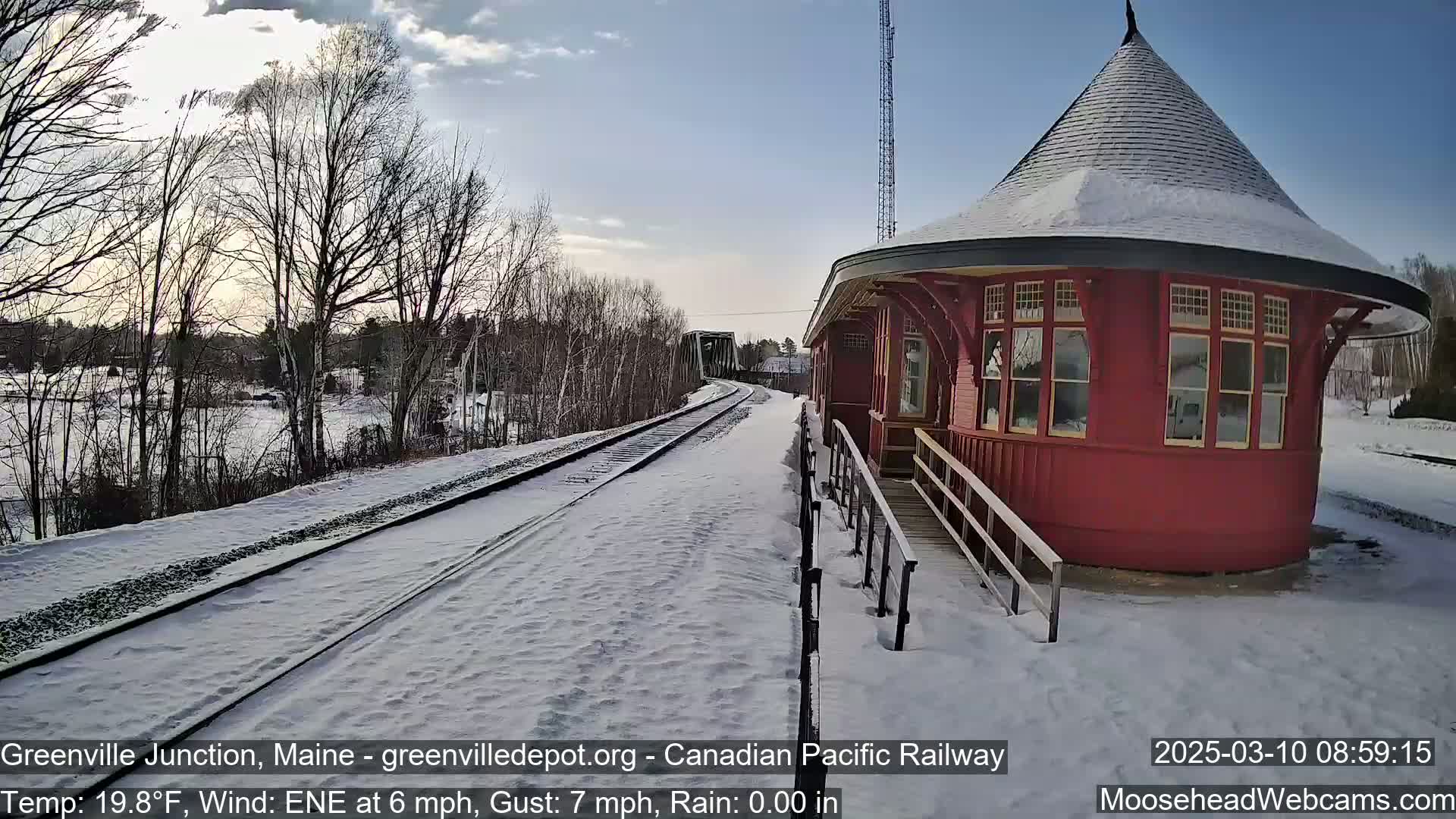 A snow-covered train platform with a red octagonal train station building is visible on a partly sunny day with a light wind.