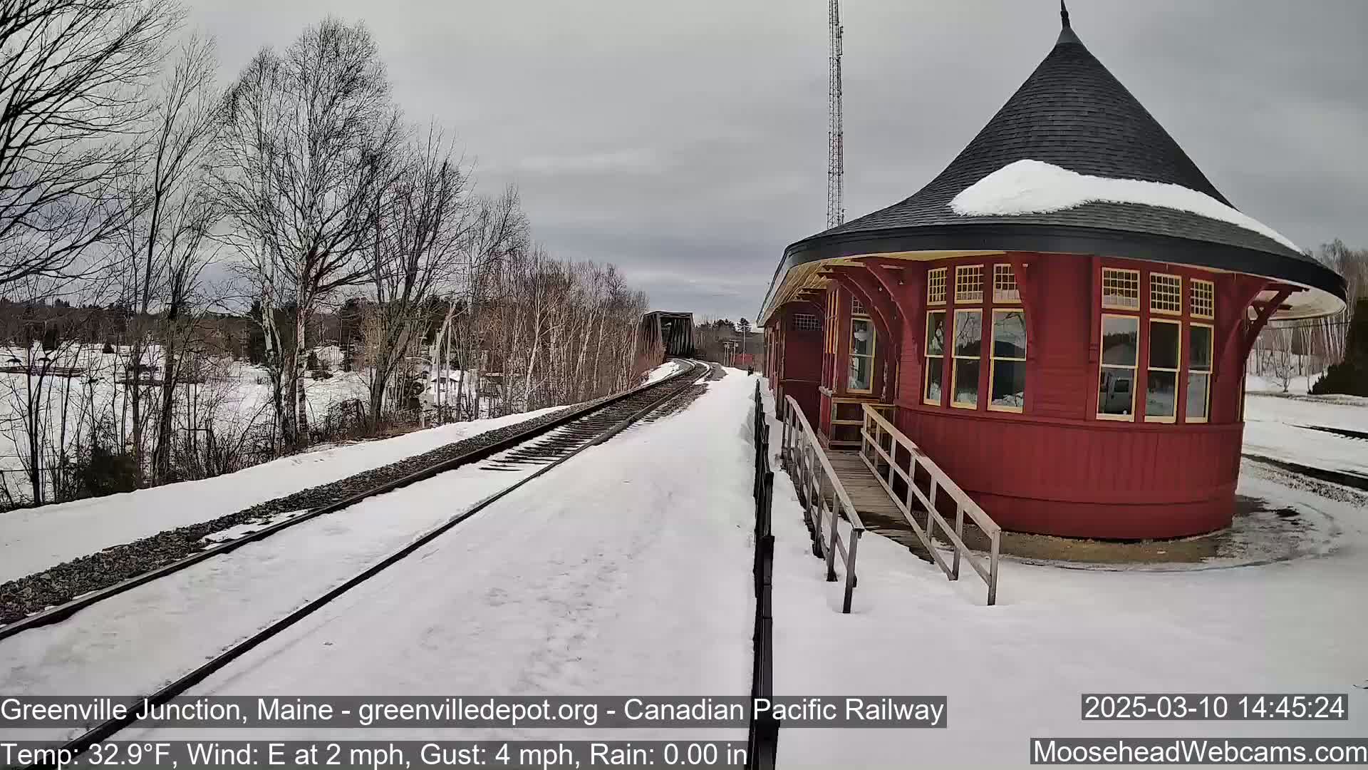 A snow-covered train platform with a red, round train station and tracks extending into the distance under a cloudy sky.