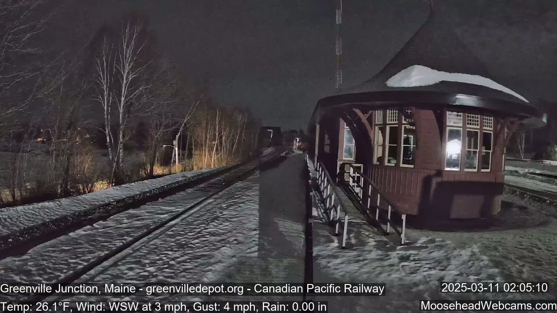 A snow-covered railway platform with a small, dark-colored building next to a set of tracks under a dark night sky.