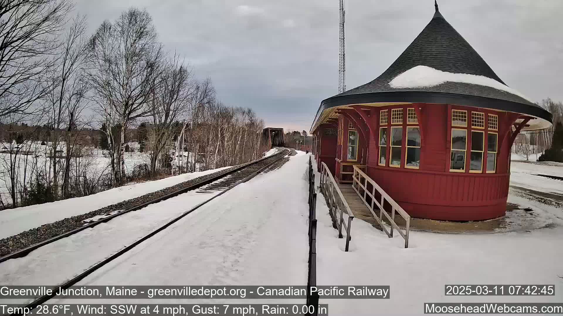A snow-covered train platform with a red, octagonal train station is visible on a cloudy day.