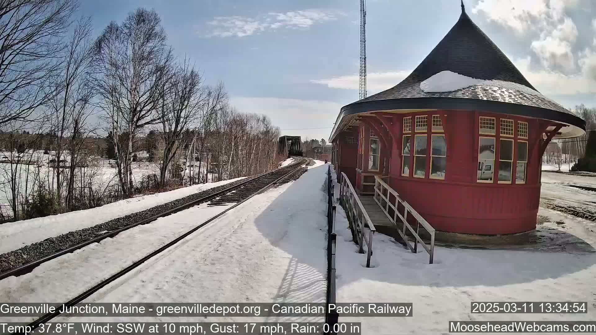 A snow-covered train platform with a red, round train depot is visible under a partly sunny sky, with a train track leading to a bridge in the distance.