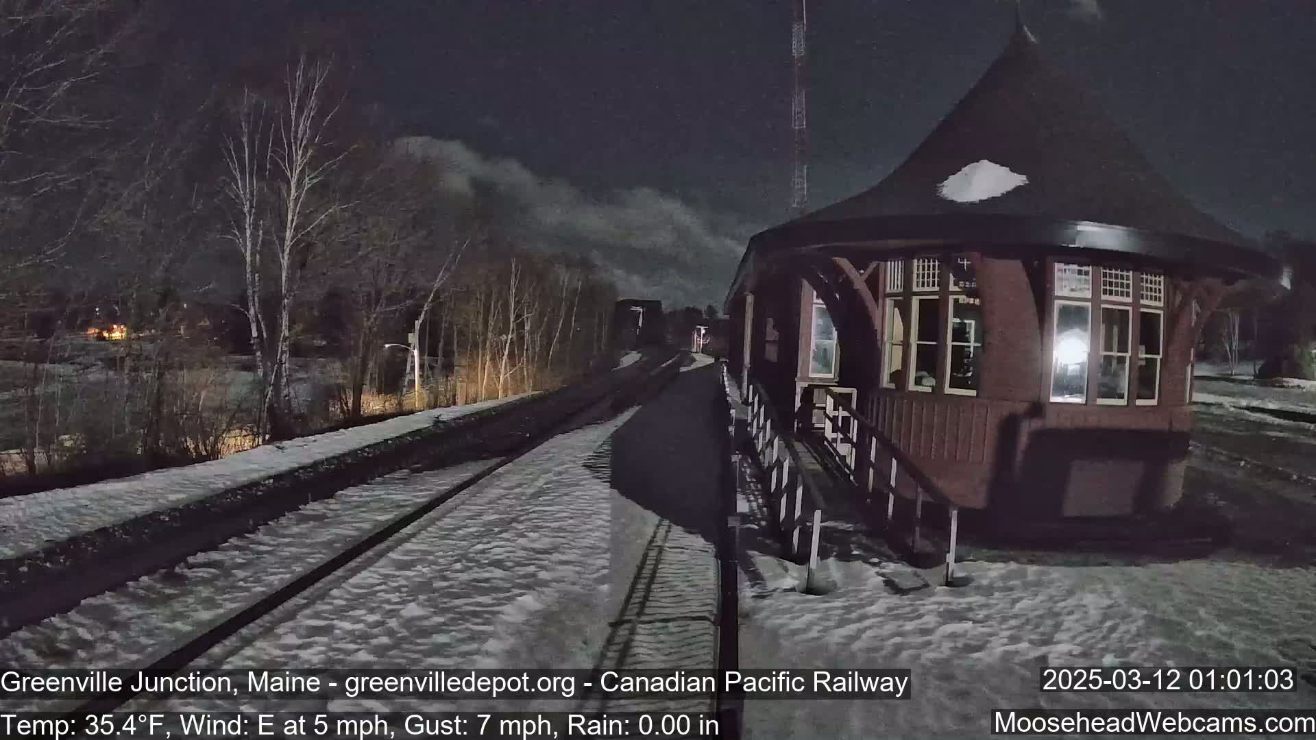 A snow-covered railway track leads to a dark brown, octagonal train station under a night sky.