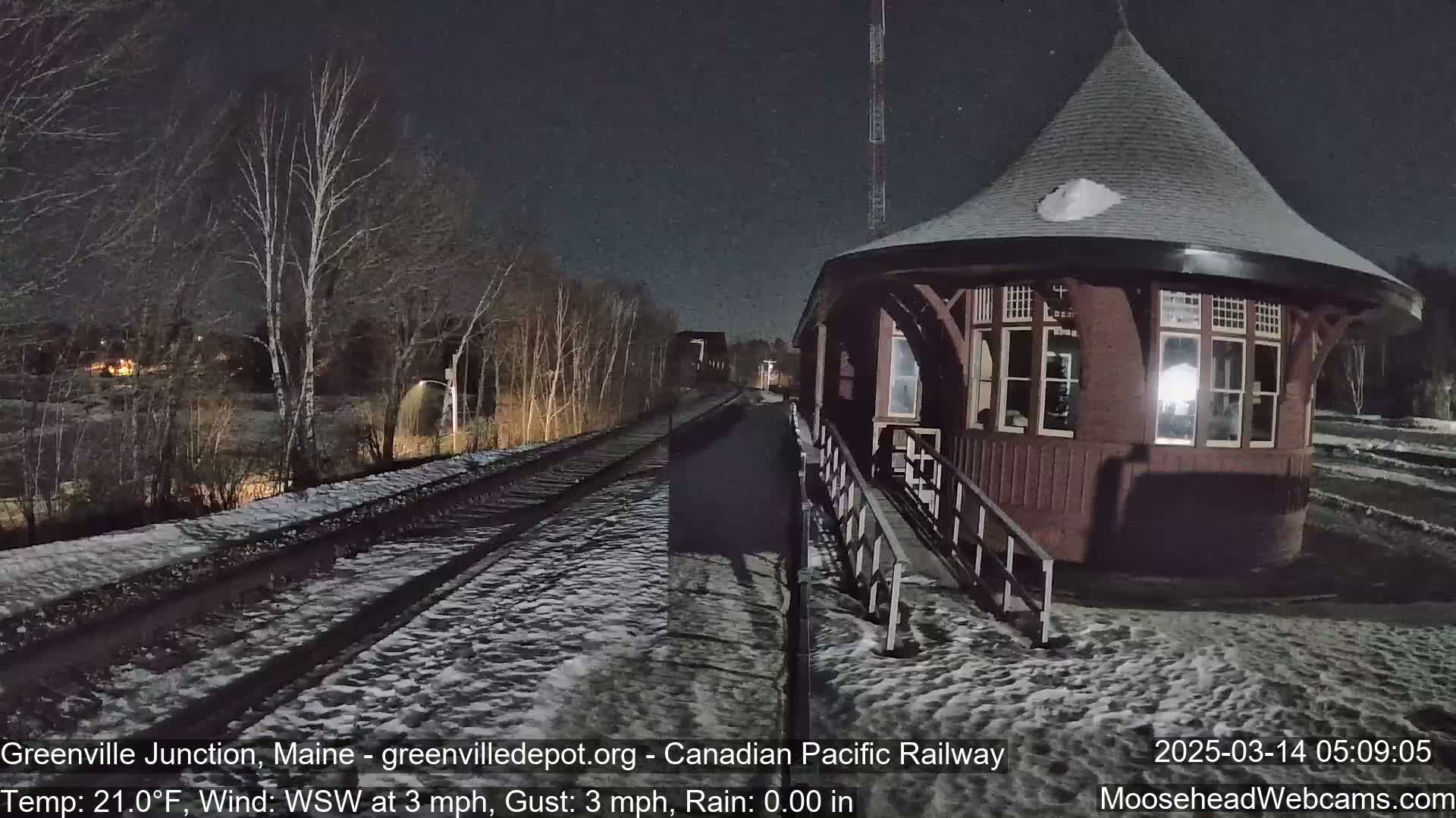 A snow-covered train platform with a red octagonal building is visible at night under a dark sky with a light wind.