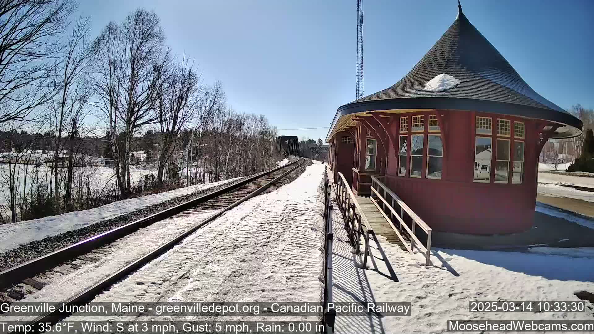 A red, octagonal train station stands beside snow-covered train tracks on a sunny day with a light south wind.